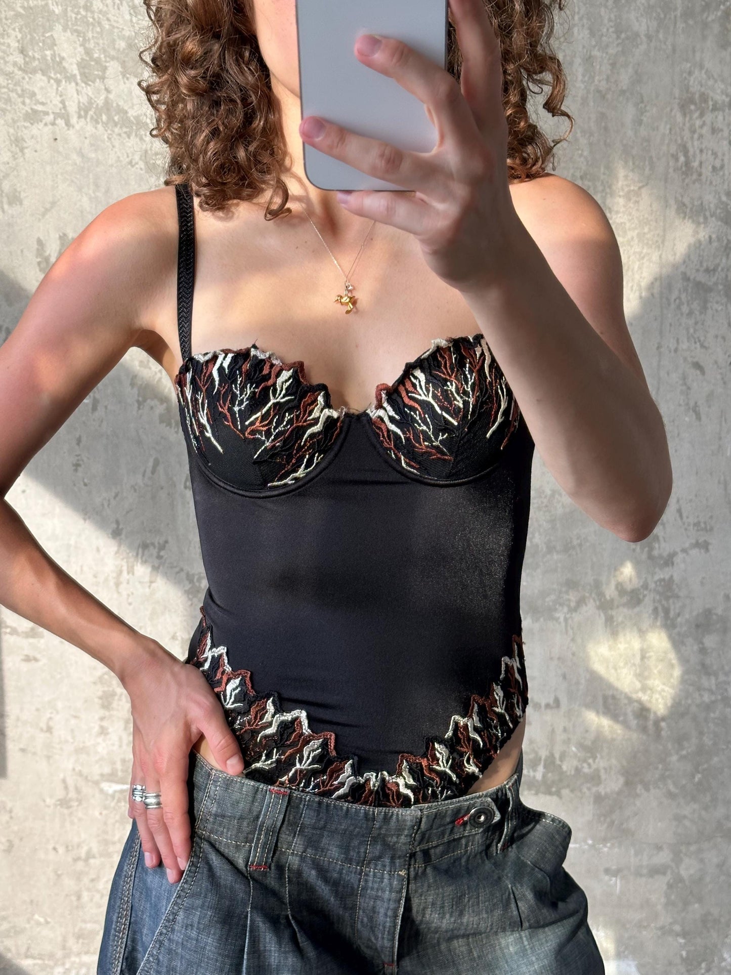 A woman is taking a selfie with a smartphone, wearing a black crop top with a floral pattern and blue jeans.