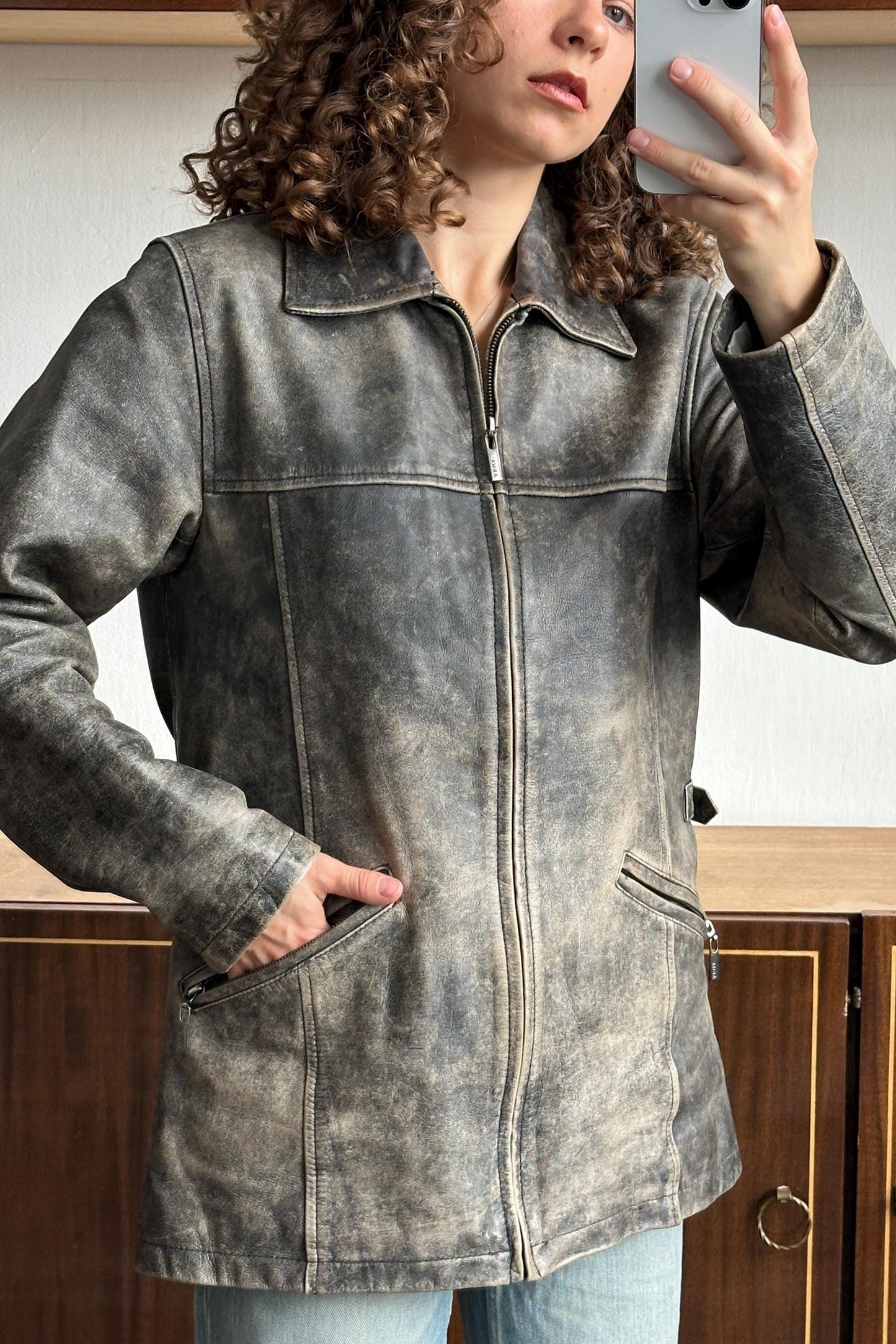 A woman with curly hair is taking a selfie with a smartphone, wearing a gray leather jacket.
