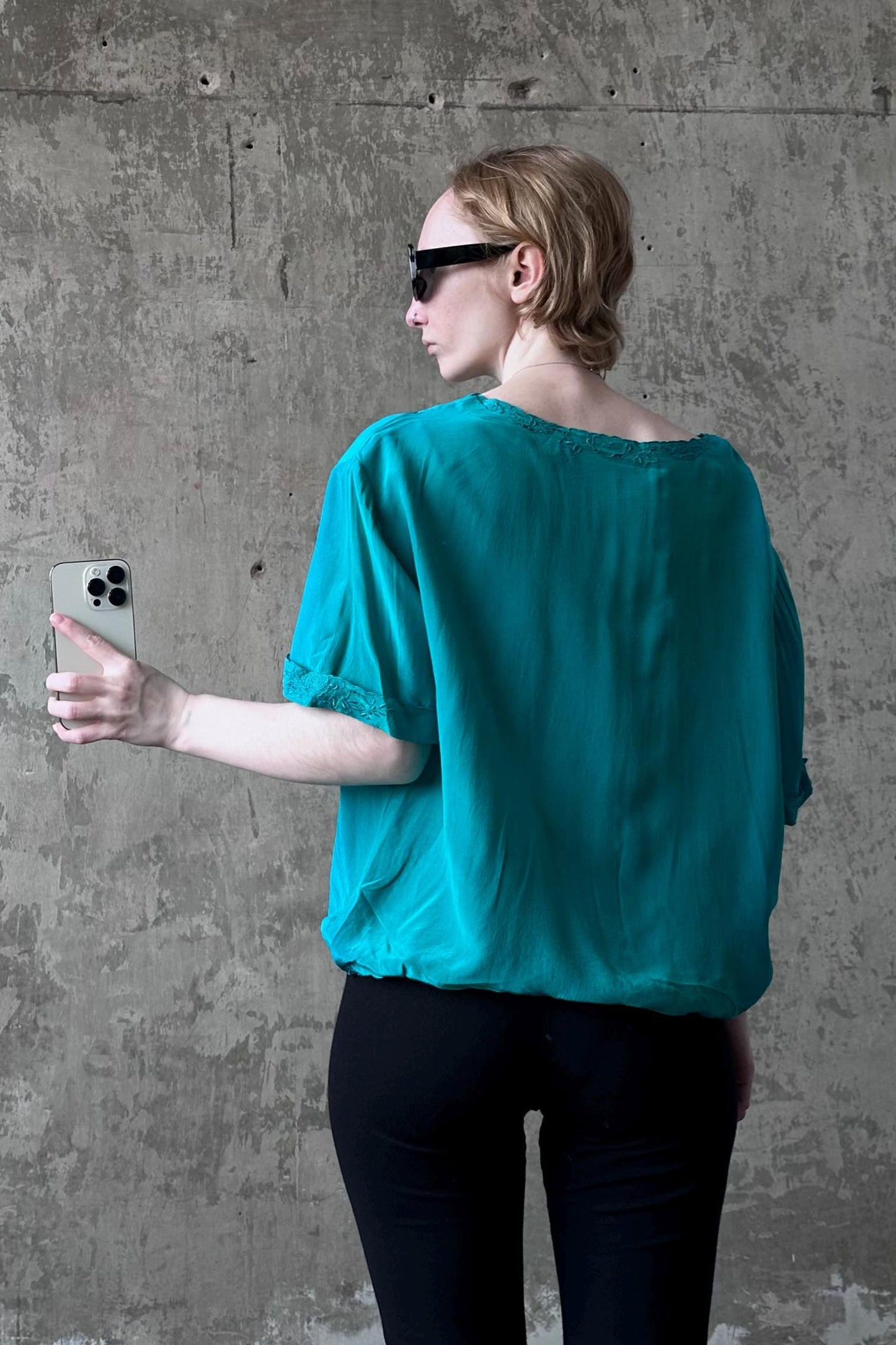 A woman in a teal blouse and sunglasses is holding a smartphone and looking at it, standing in front of a concrete wall.