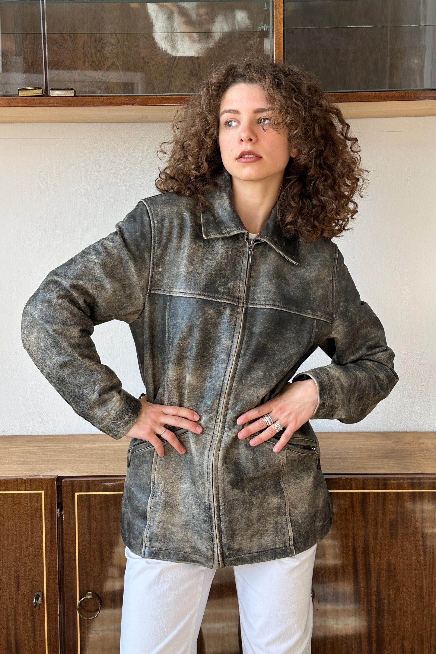 A woman with curly hair wearing a gray leather jacket stands in front of a wooden cabinet.