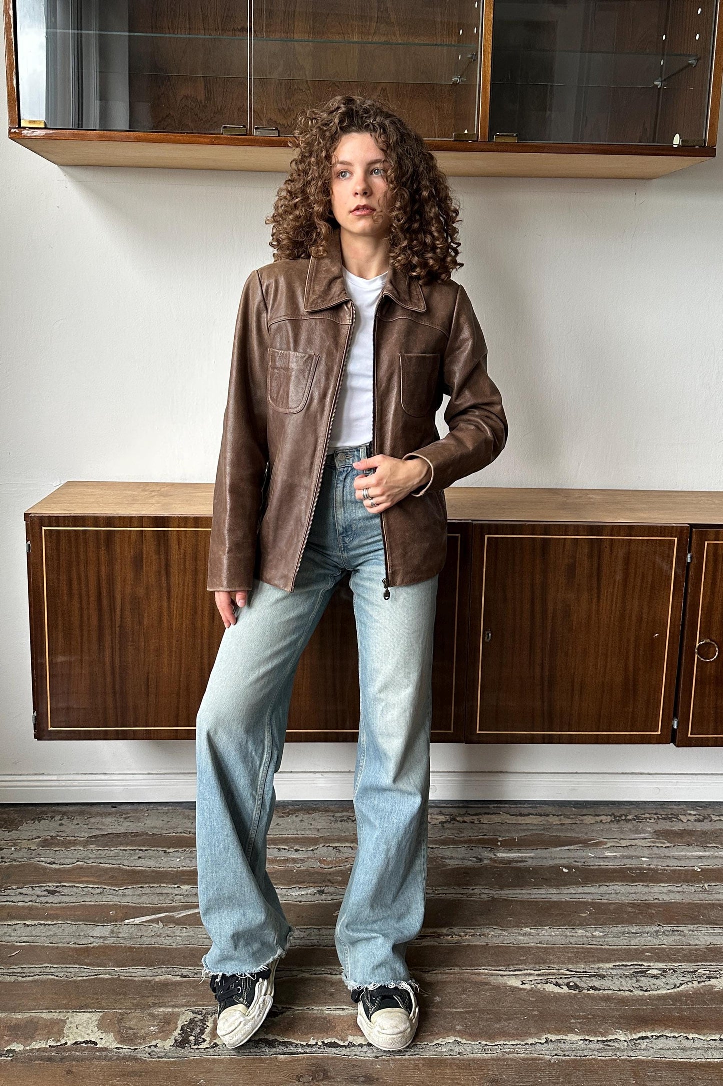 A woman with curly hair wearing a brown leather jacket, white shirt, and blue jeans stands in a room with wooden cabinets.