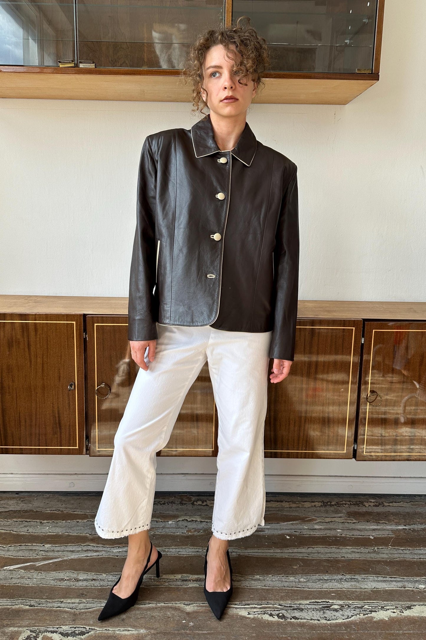 A woman wearing a black leather jacket and white pants stands in front of a wooden cabinet.