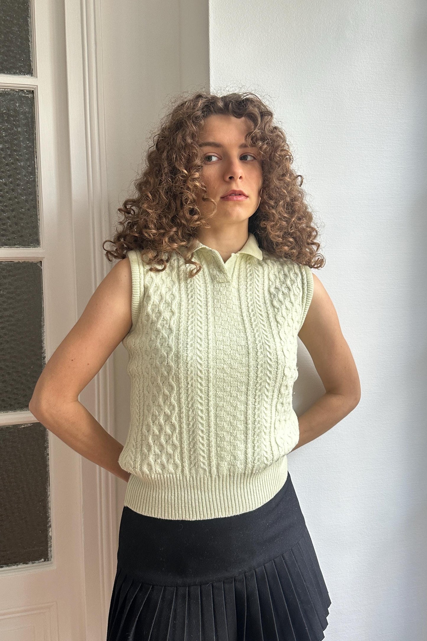 A young woman with curly hair stands in front of a white wall, wearing a sleeveless white sweater and a black skirt.
