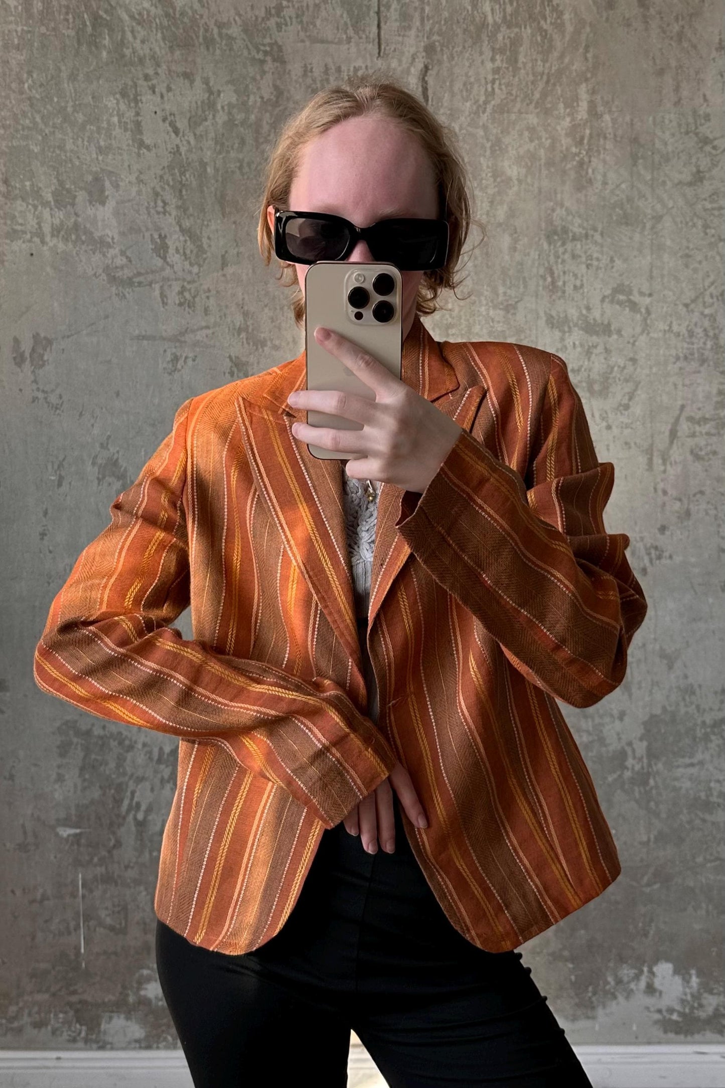 A person wearing a striped orange blazer and sunglasses is taking a selfie with a smartphone.
