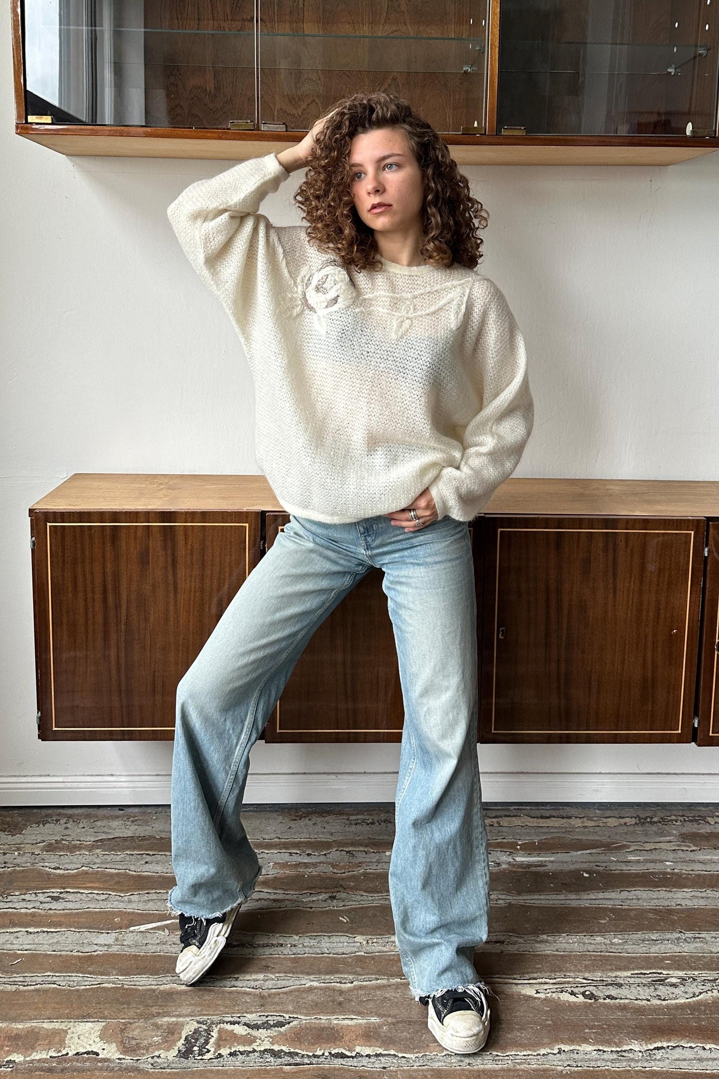 A woman with curly hair wearing a white sweater and blue jeans stands in a room with wooden cabinets in the background.