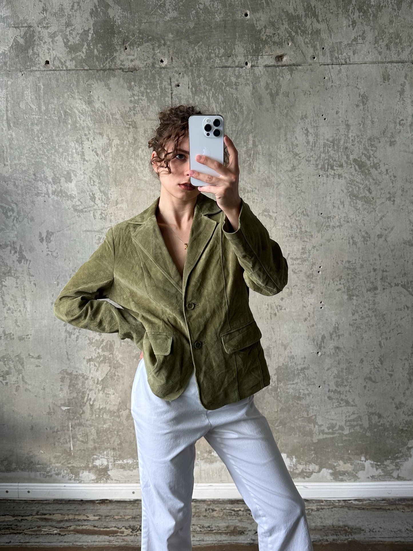 A person is taking a selfie with a white iPhone, wearing a green jacket and white pants, against a concrete wall background.