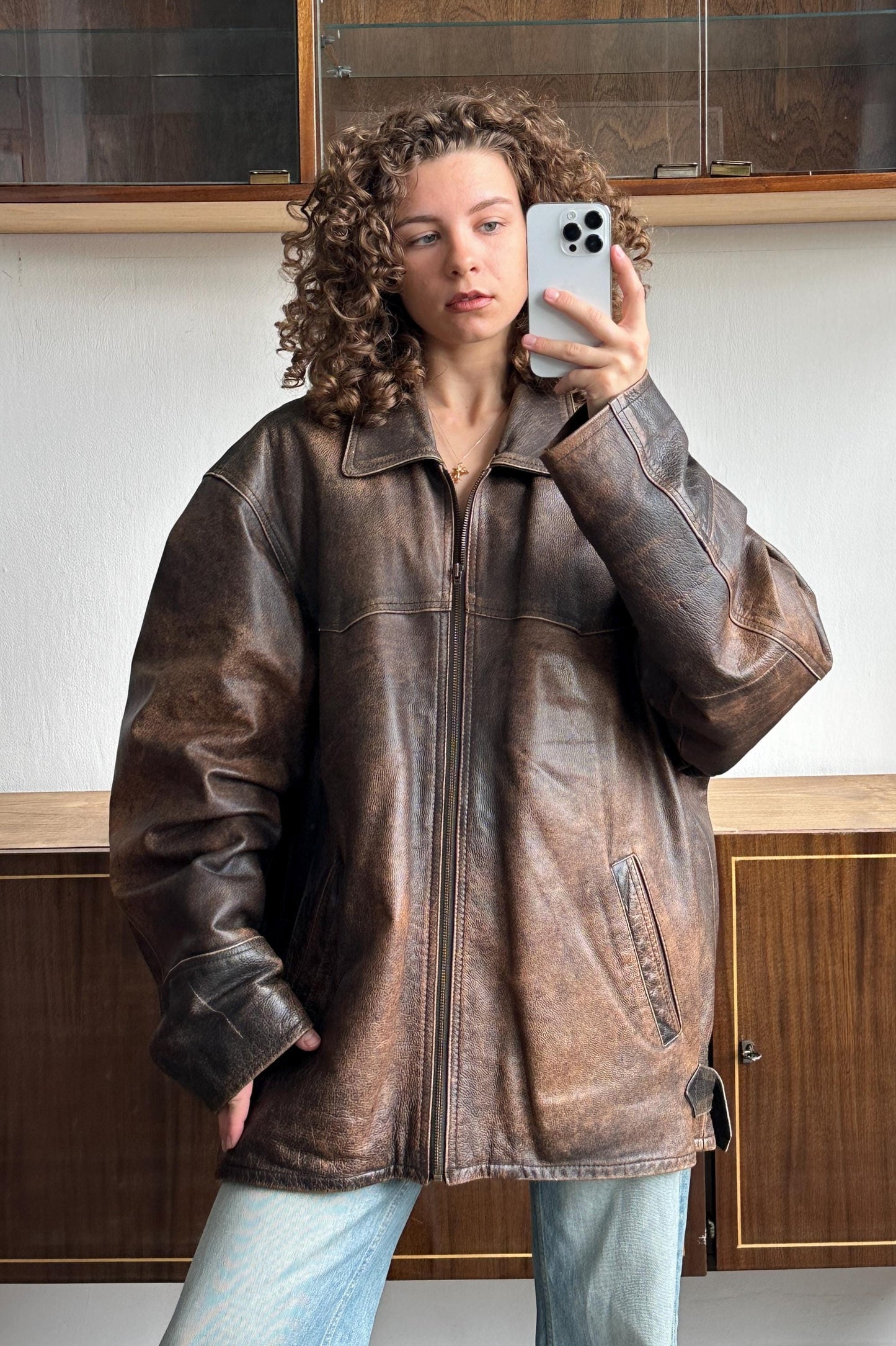 A woman with curly hair is taking a selfie with a smartphone, wearing a brown leather jacket.