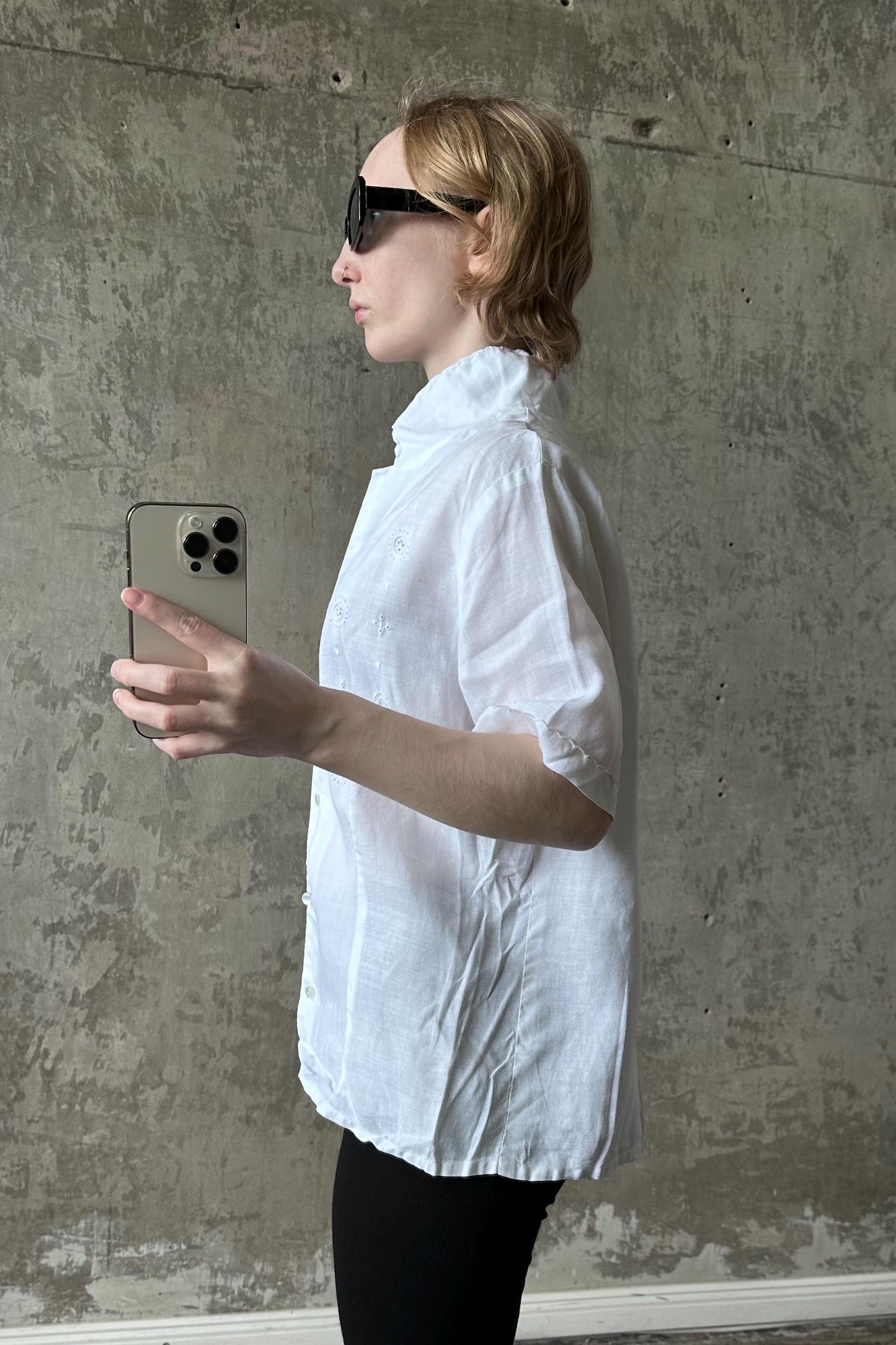 A person wearing a white shirt and sunglasses is holding a smartphone and appears to be taking a selfie in front of a concrete wall.