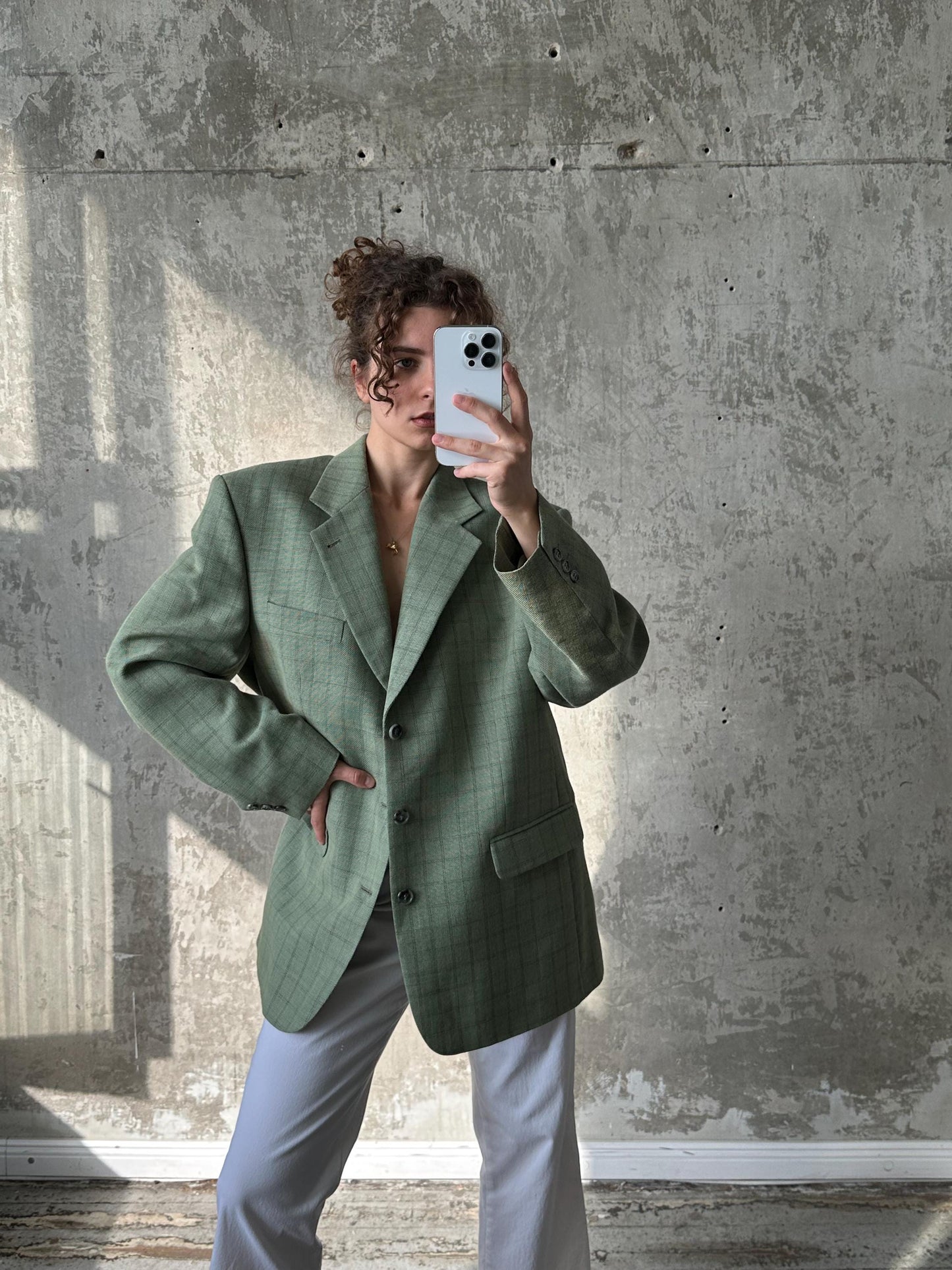 A person is taking a selfie with a white phone, wearing a green blazer and standing in front of a concrete wall.
