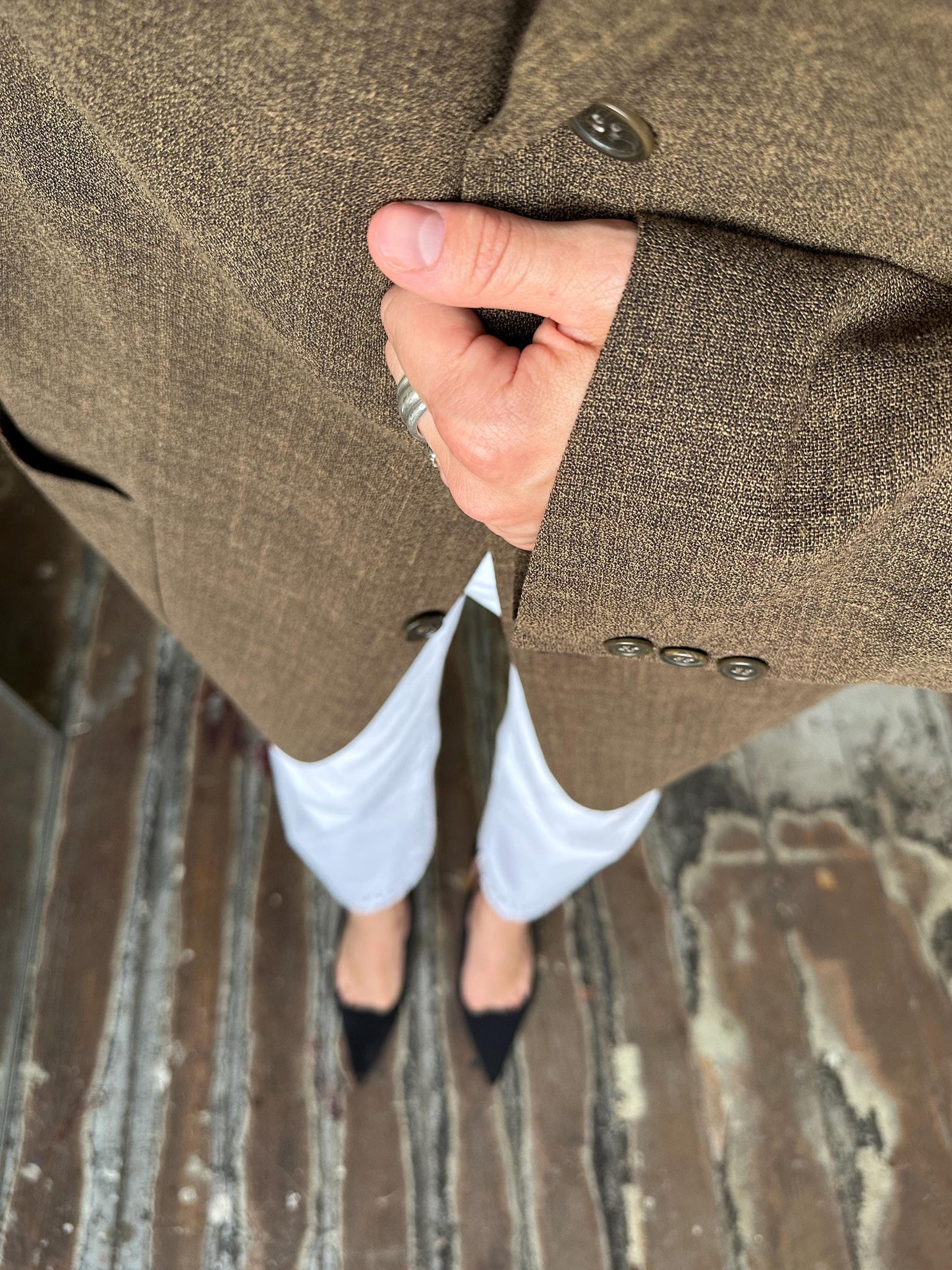 A person wearing a brown tweed jacket and white pants stands on a wooden floor, with their hand resting on their jacket.
