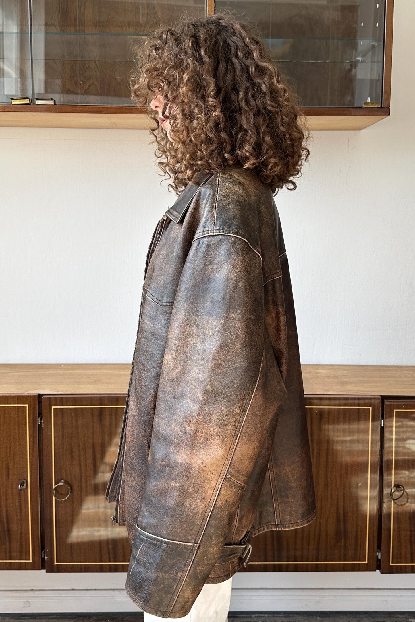 A person with curly hair wearing a brown leather jacket stands in front of a wooden cabinet.