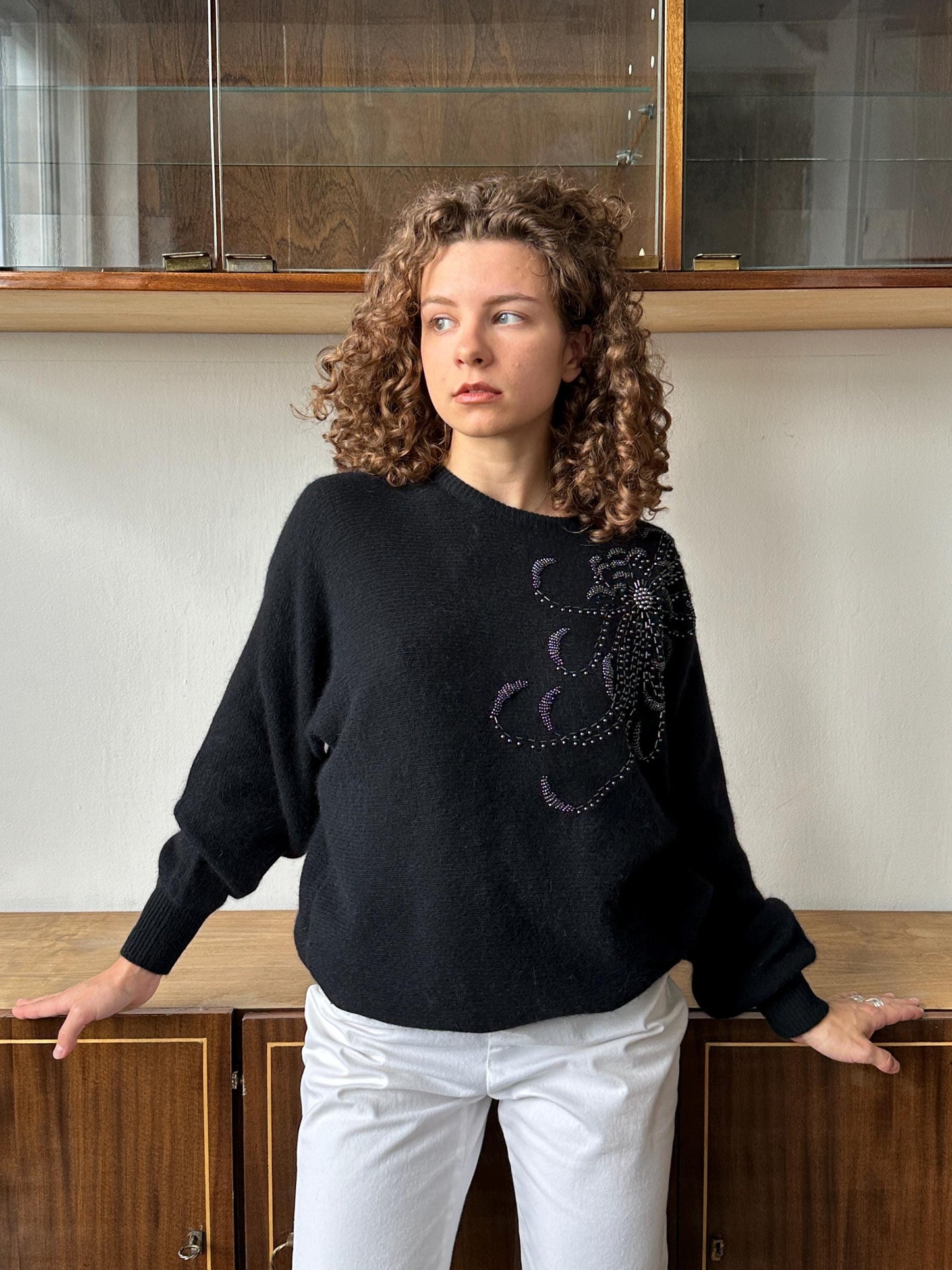A woman with curly hair wearing a black sweater with purple designs stands in front of a wooden cabinet.