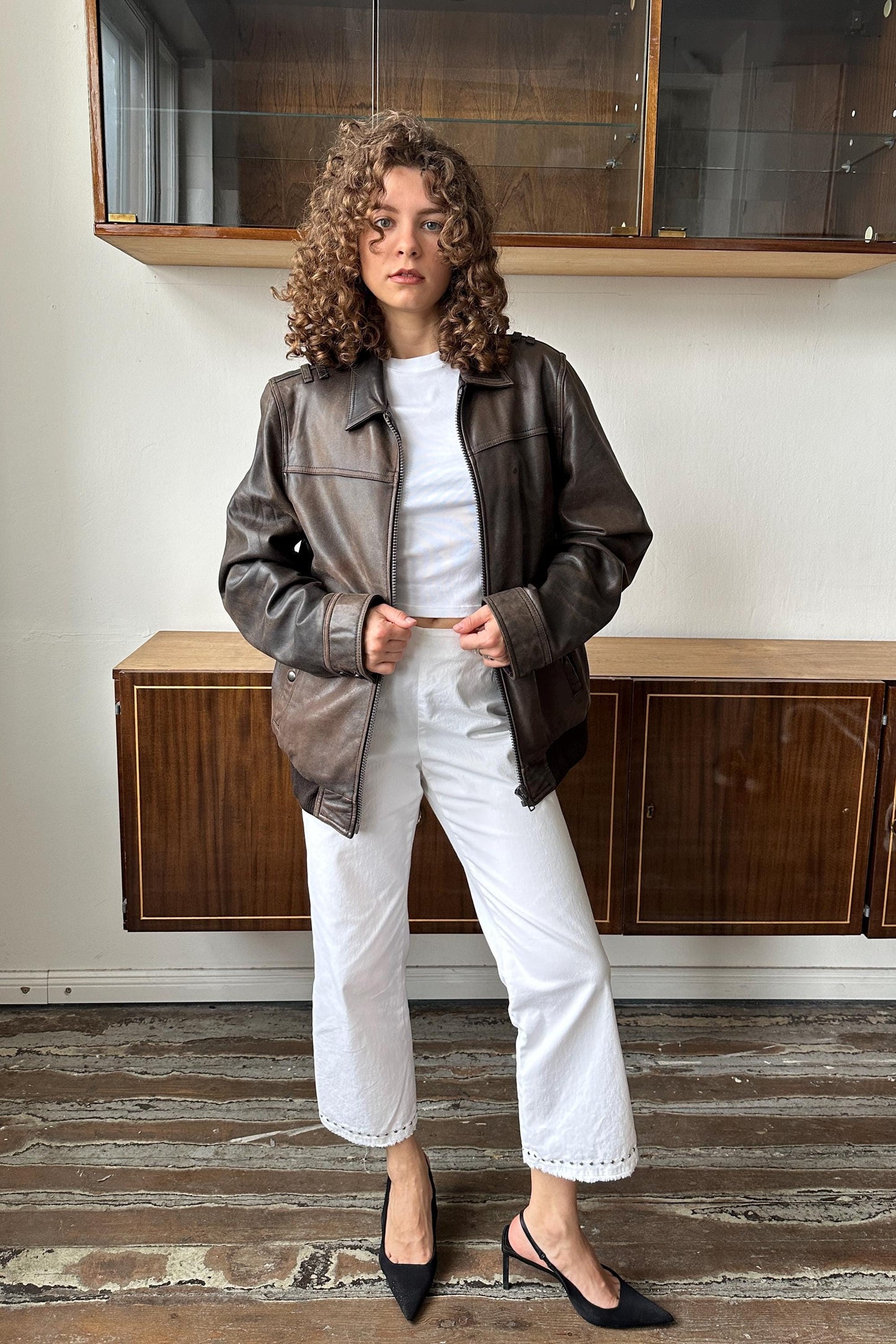 A woman with curly hair wearing a brown leather jacket, white pants, and black heels stands in a room with wooden cabinets.