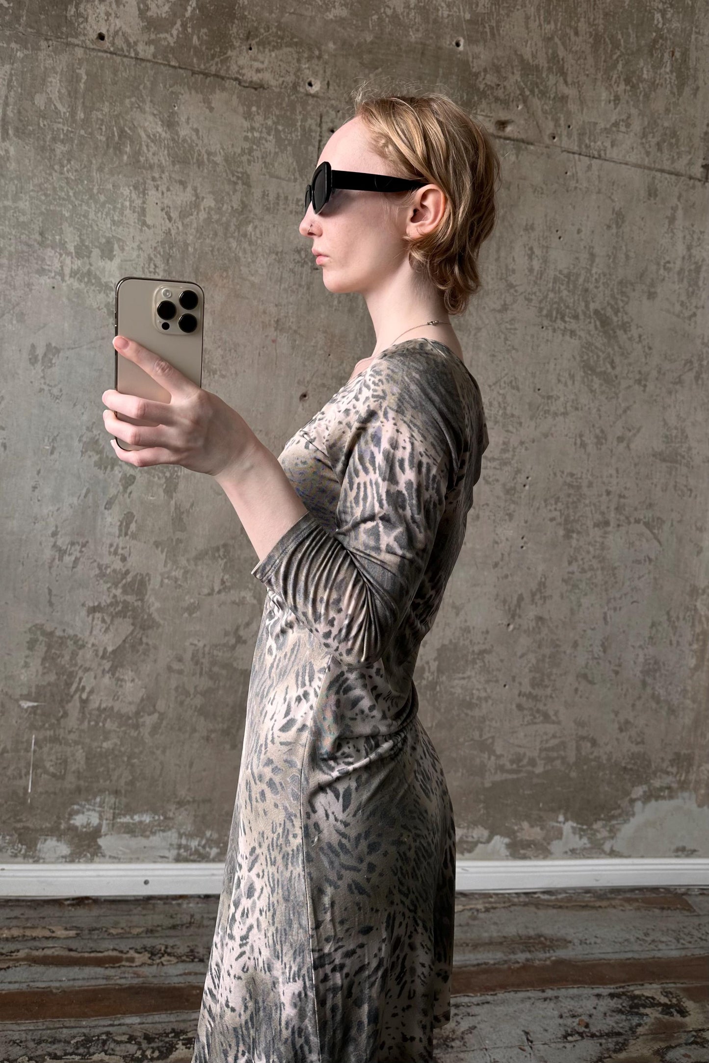 A woman wearing a leopard print dress and sunglasses is holding a smartphone and taking a selfie in front of a concrete wall.