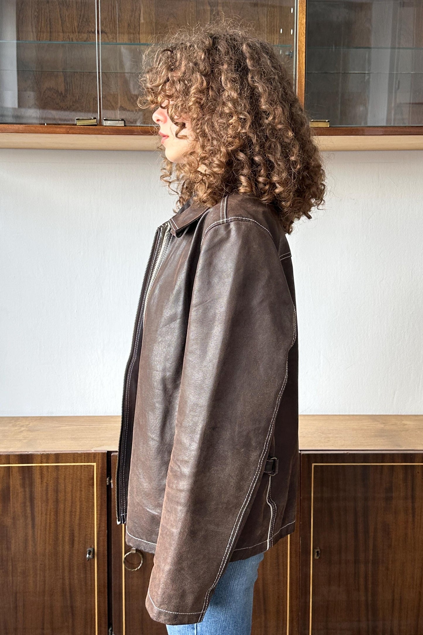 A woman with curly hair wearing a brown leather jacket stands in front of a wooden cabinet.