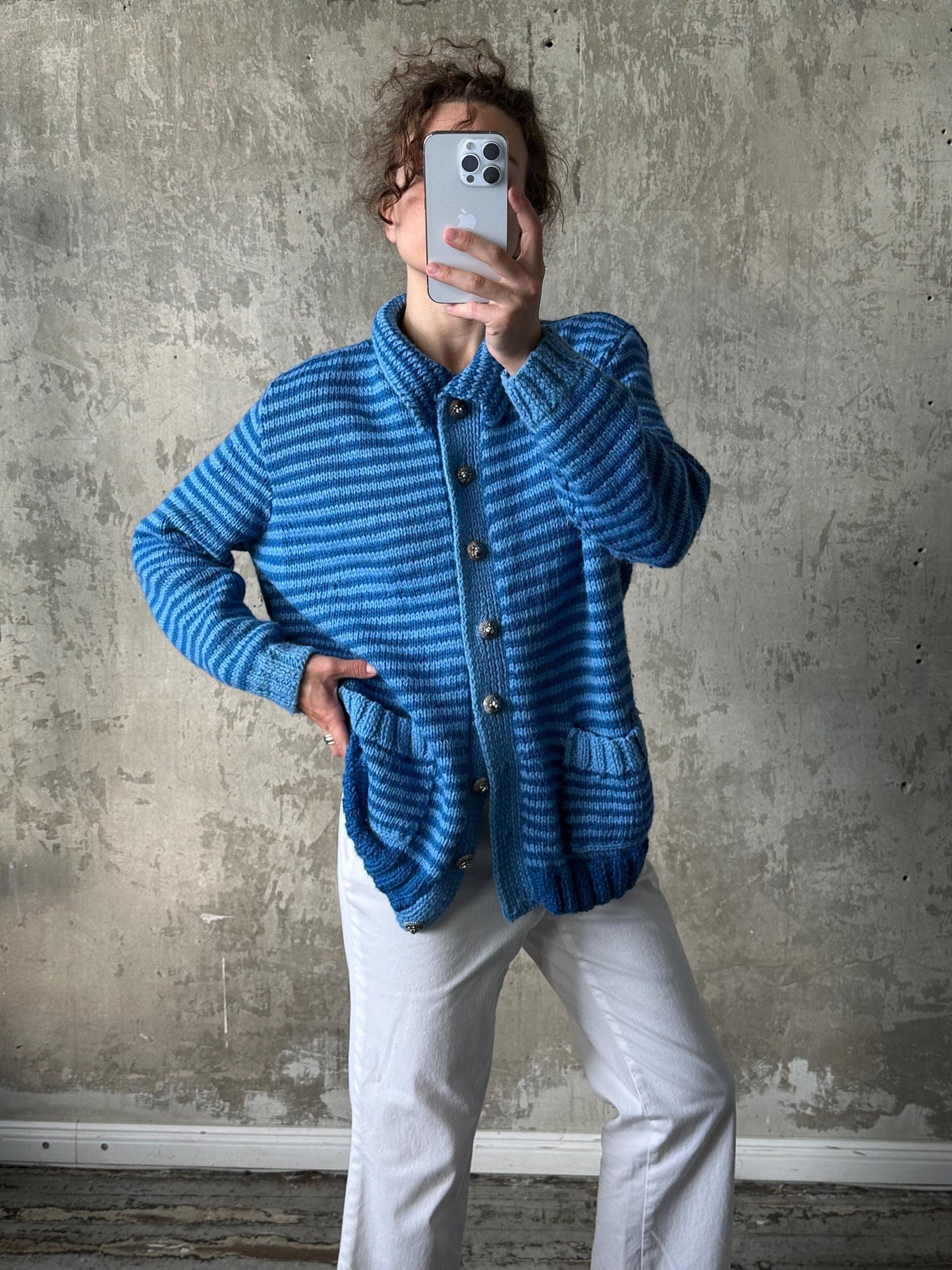 A person is taking a selfie with a striped blue cardigan and white pants, holding a smartphone up to their face.