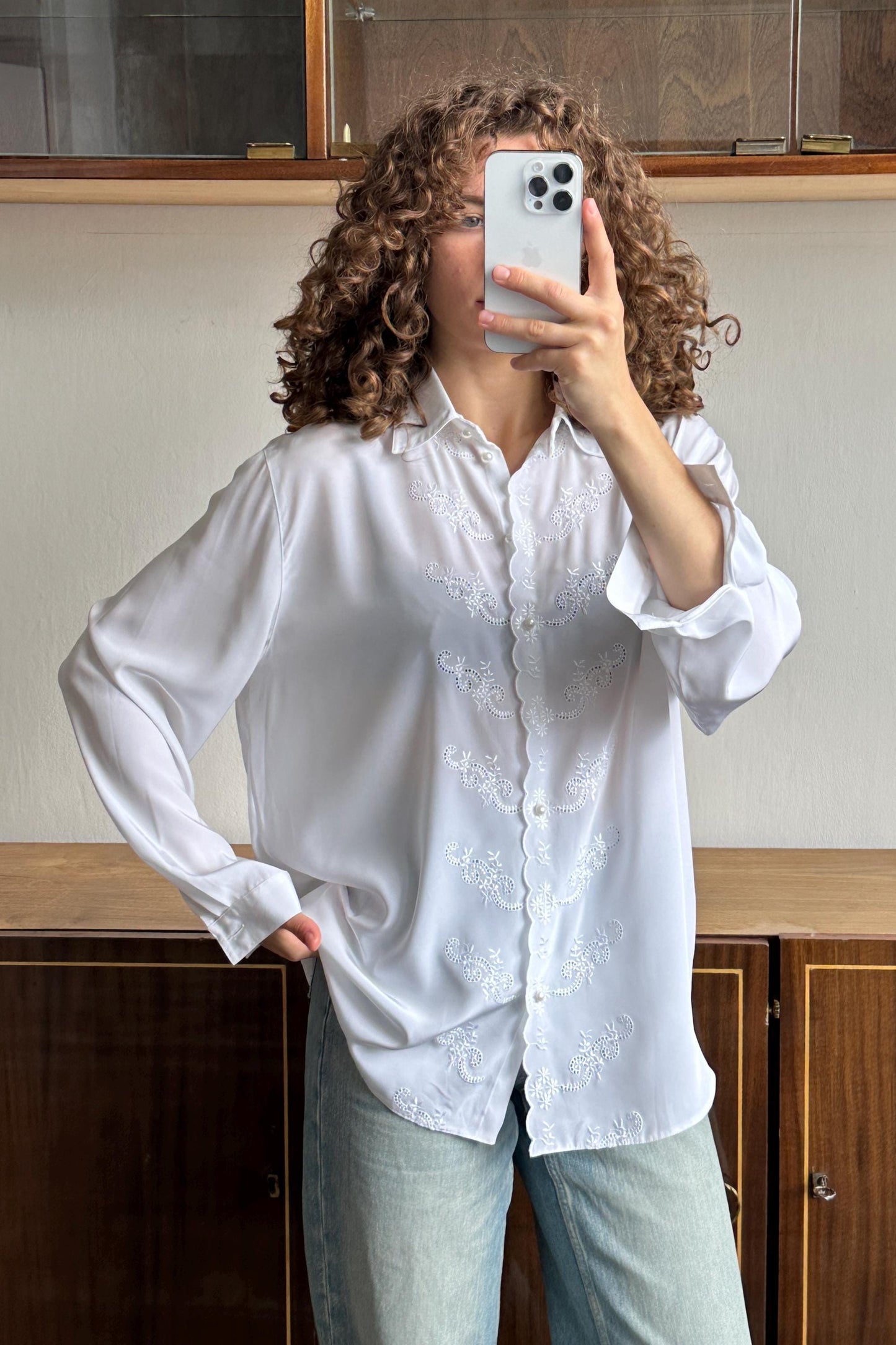A woman with curly hair is taking a selfie with a white smartphone, wearing a white blouse with embroidered details.