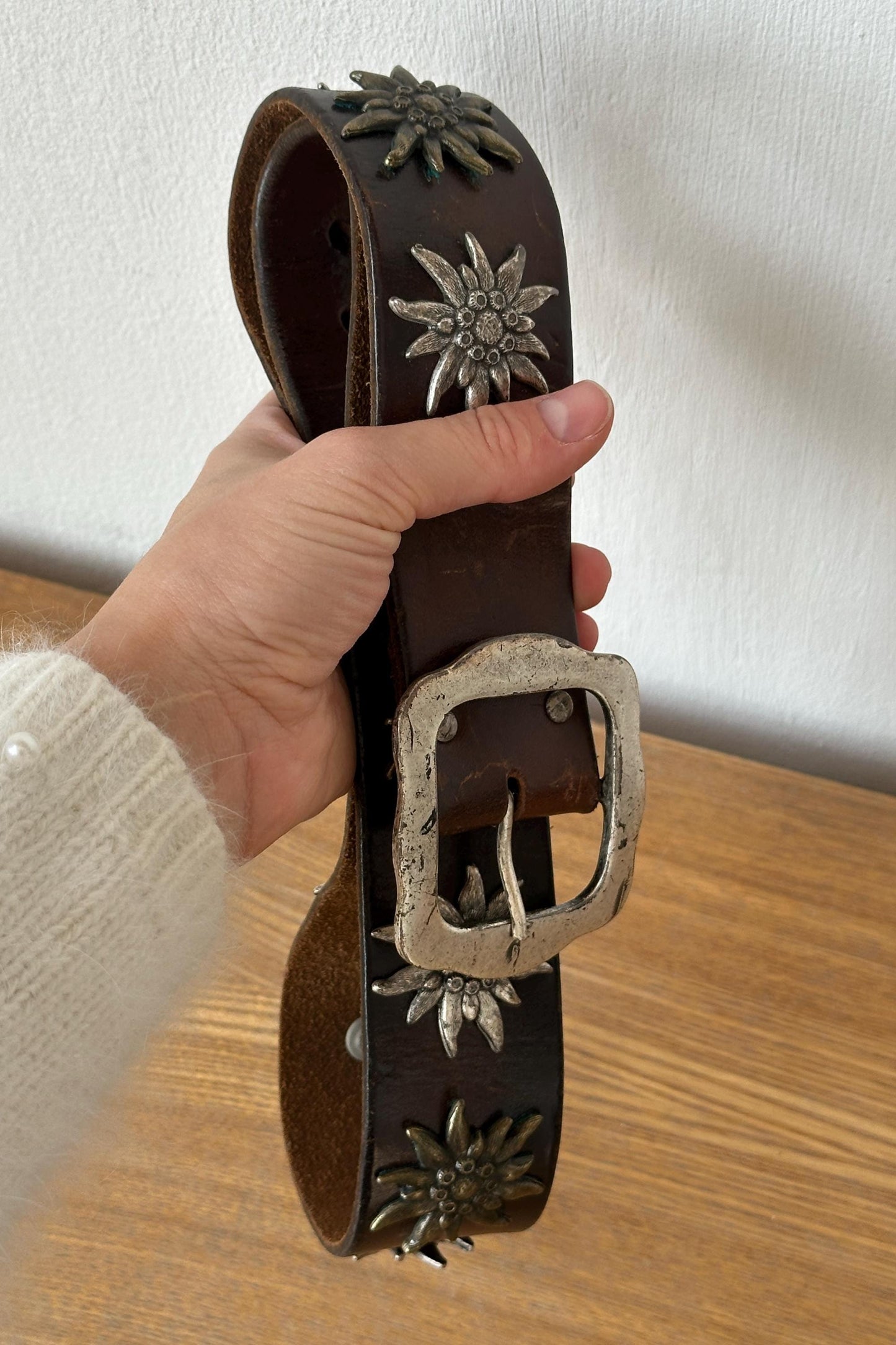 A hand is holding a brown leather belt with metal studs and a large metal buckle.