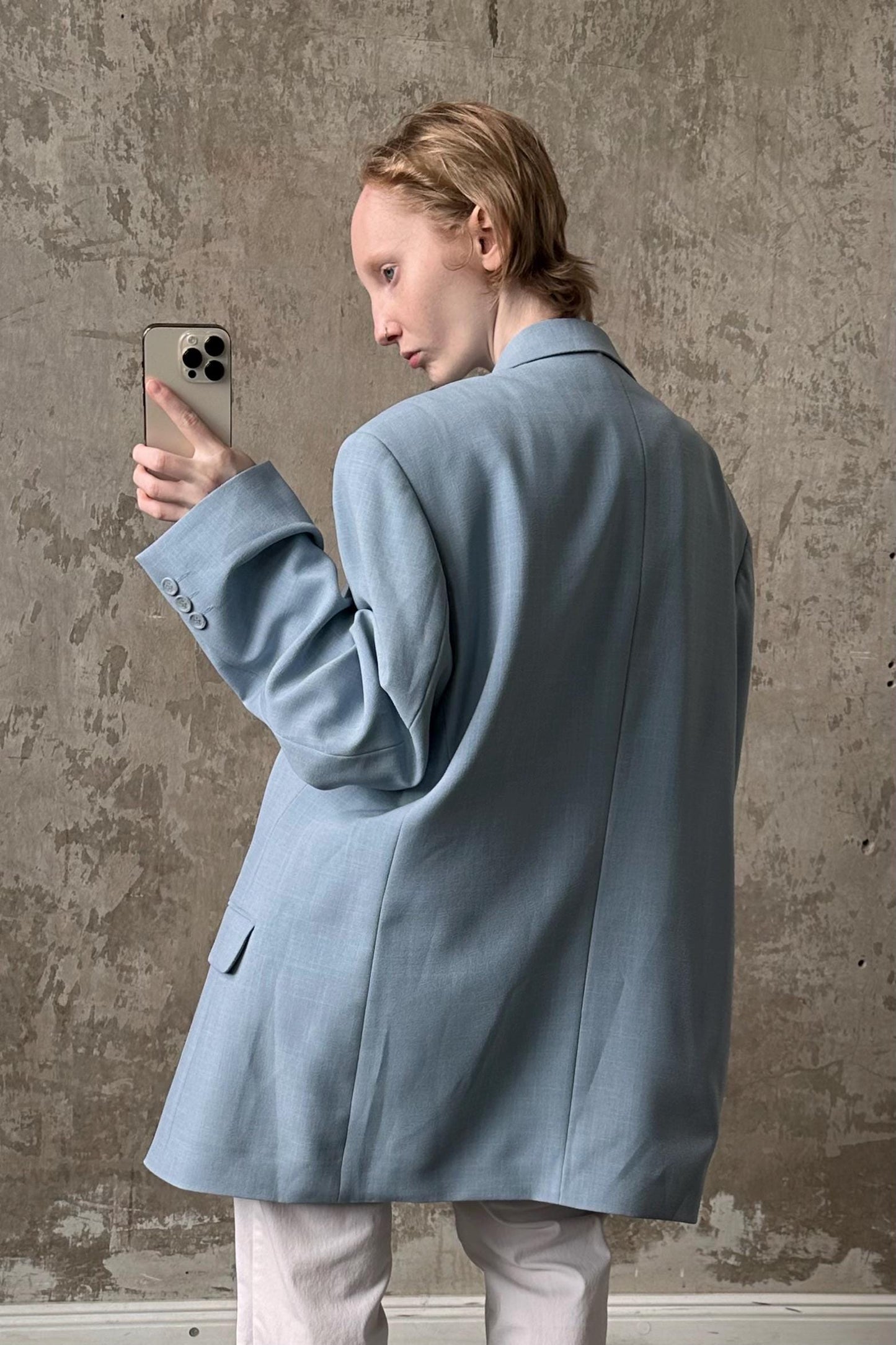 A woman in a light blue blazer is holding a smartphone and looking at it, with a neutral expression on her face.