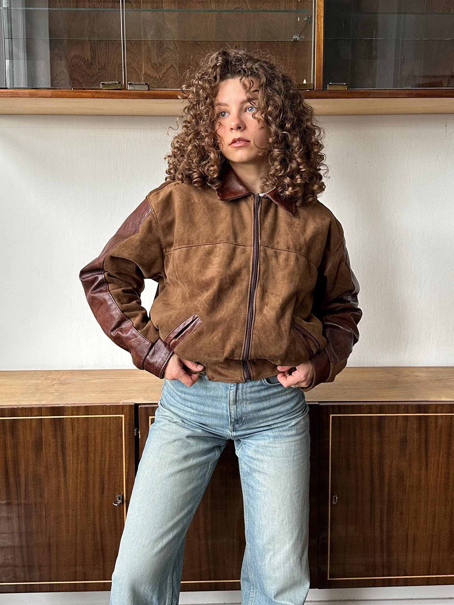 A person with curly hair wearing a brown jacket and blue jeans stands in a room with wooden cabinets in the background.