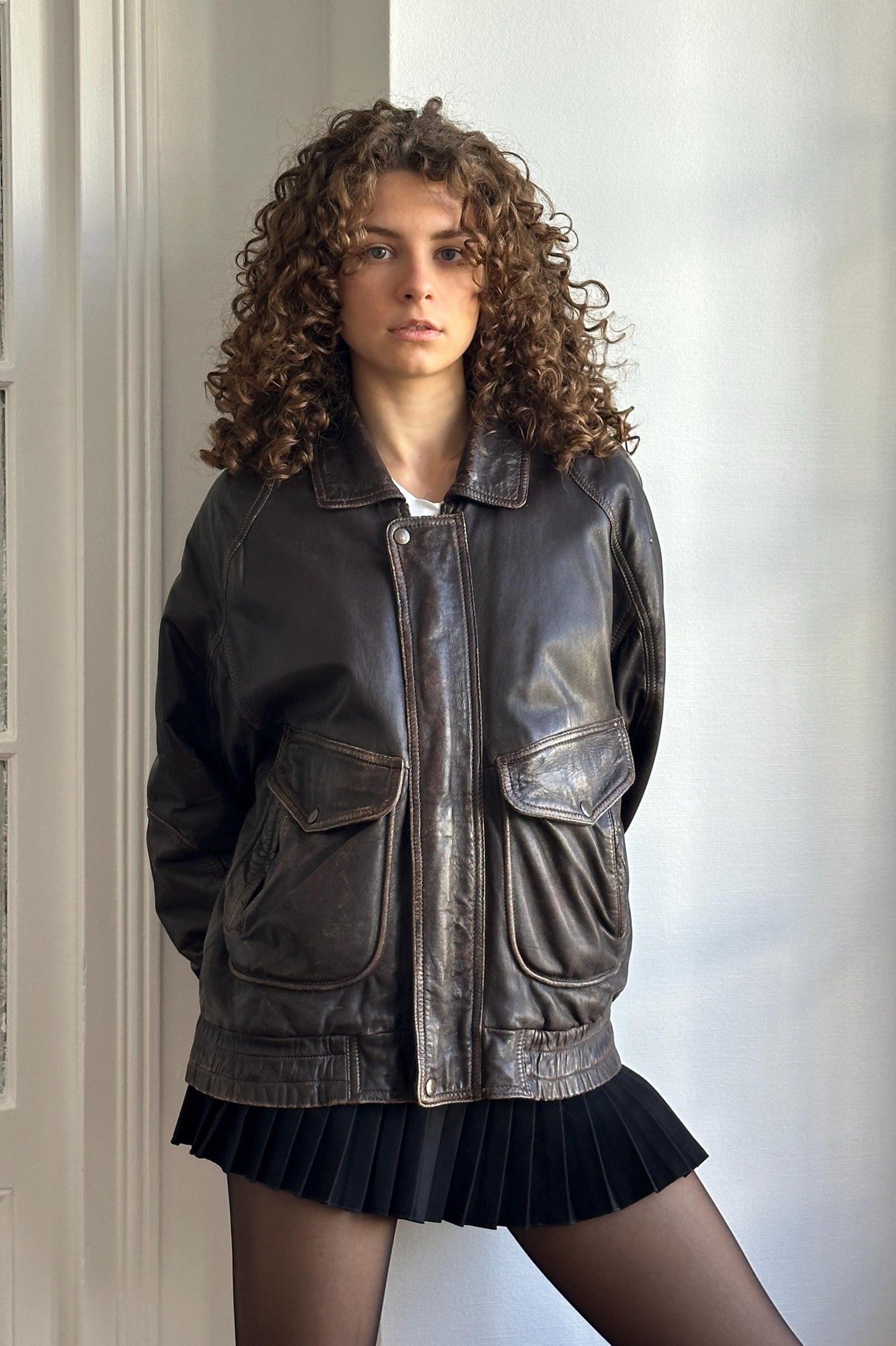 A young woman with curly hair wearing a black leather jacket and a black skirt stands in front of a white wall.