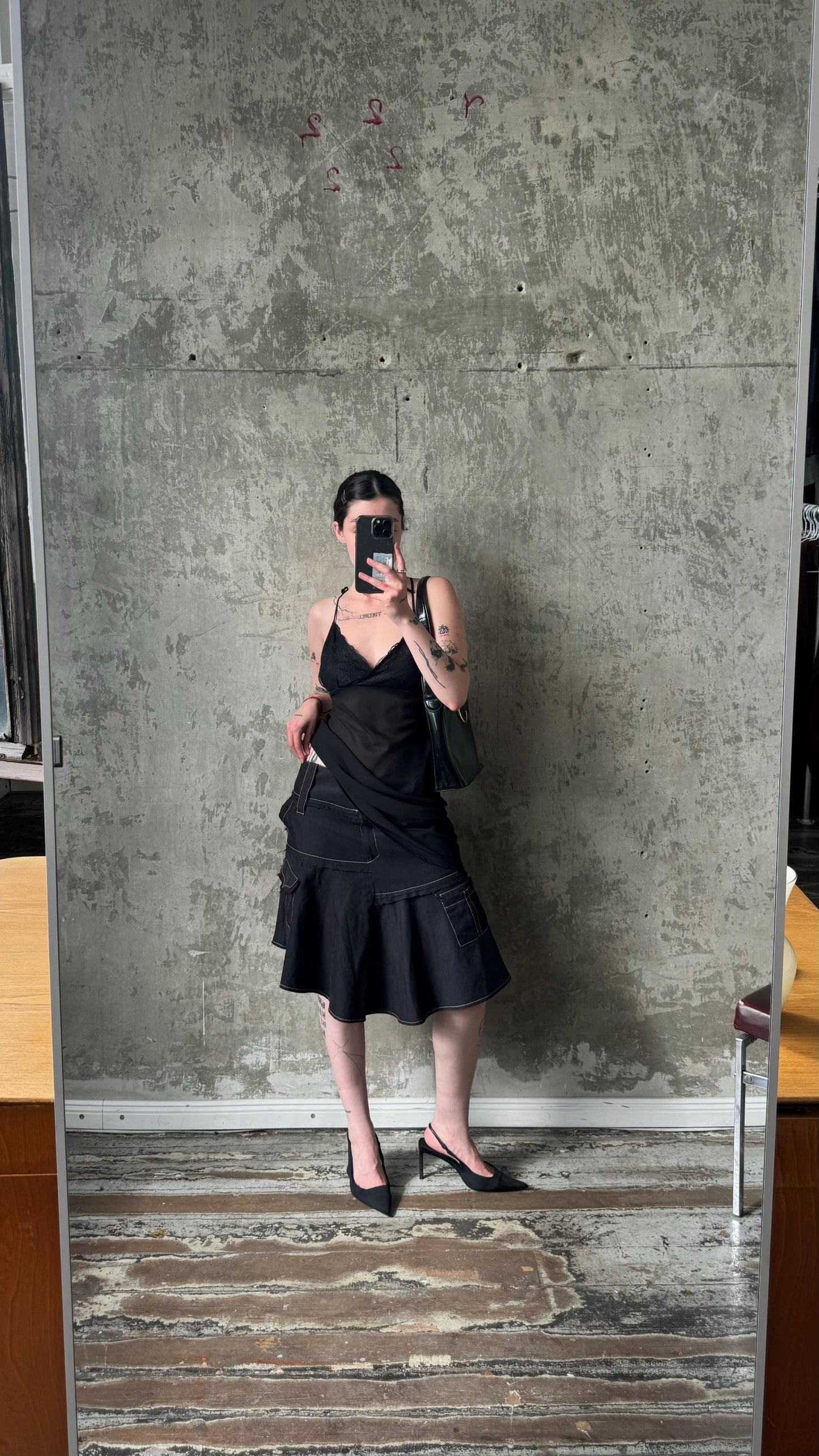 A person is taking a selfie in front of a concrete wall, holding a phone and wearing a black dress and heels.