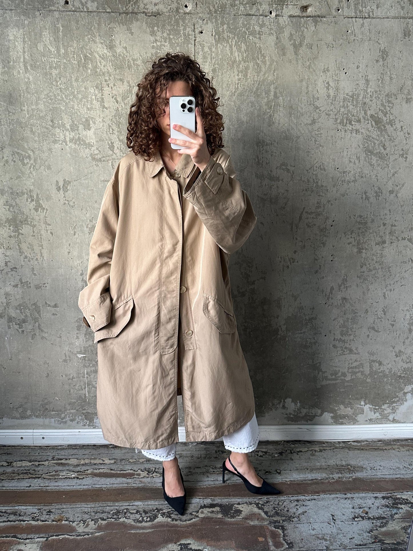 A person is taking a selfie with a smartphone, wearing a beige trench coat and black heels, against a concrete wall.