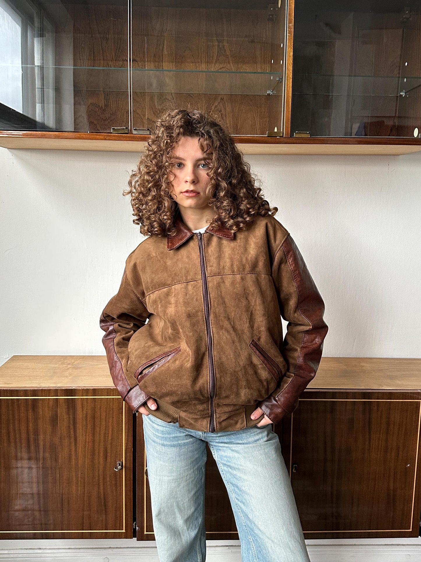A person with curly hair wearing a brown leather jacket and blue jeans stands in front of a wooden cabinet.