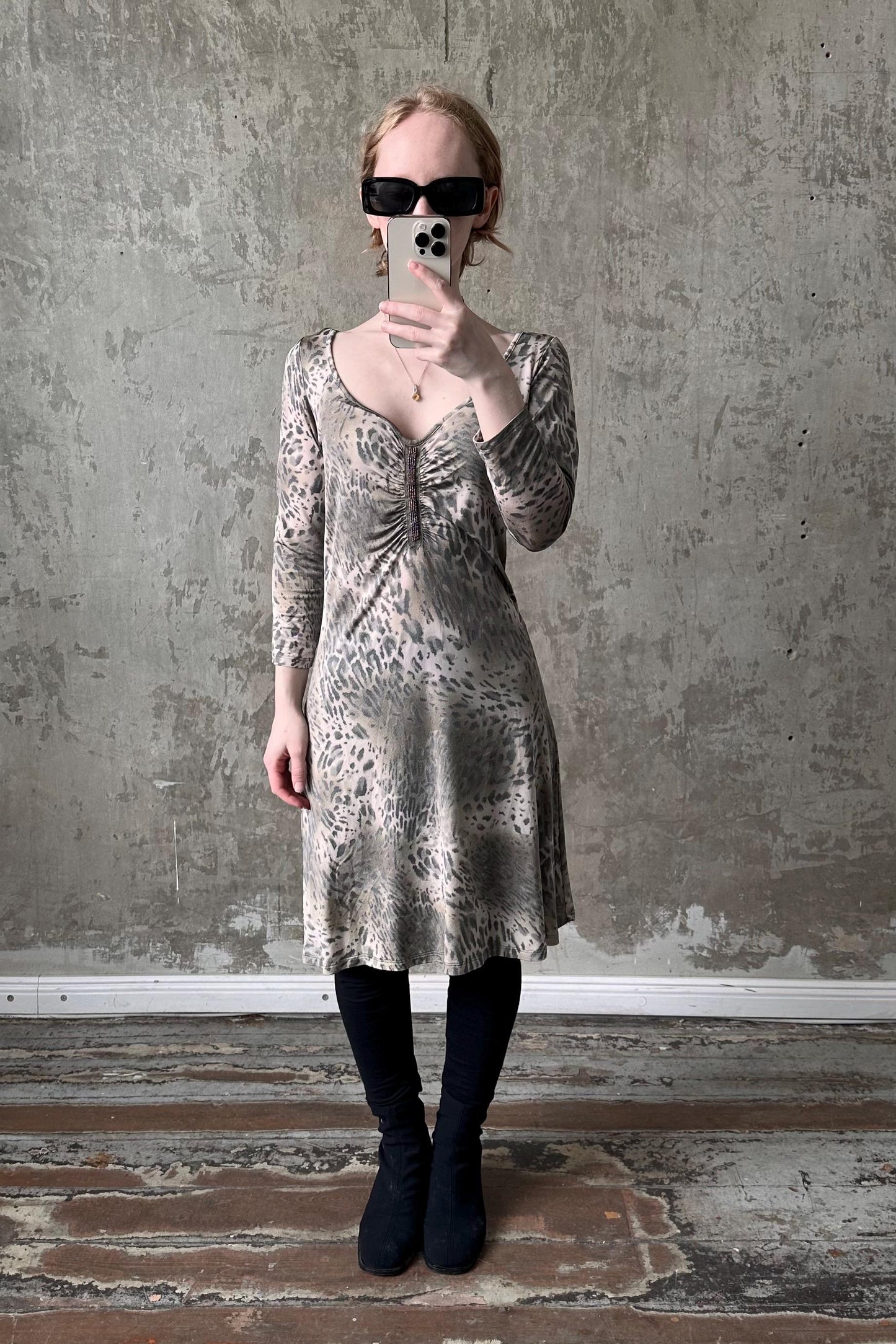 A woman wearing a leopard print dress and sunglasses is taking a selfie with her phone against a concrete wall.