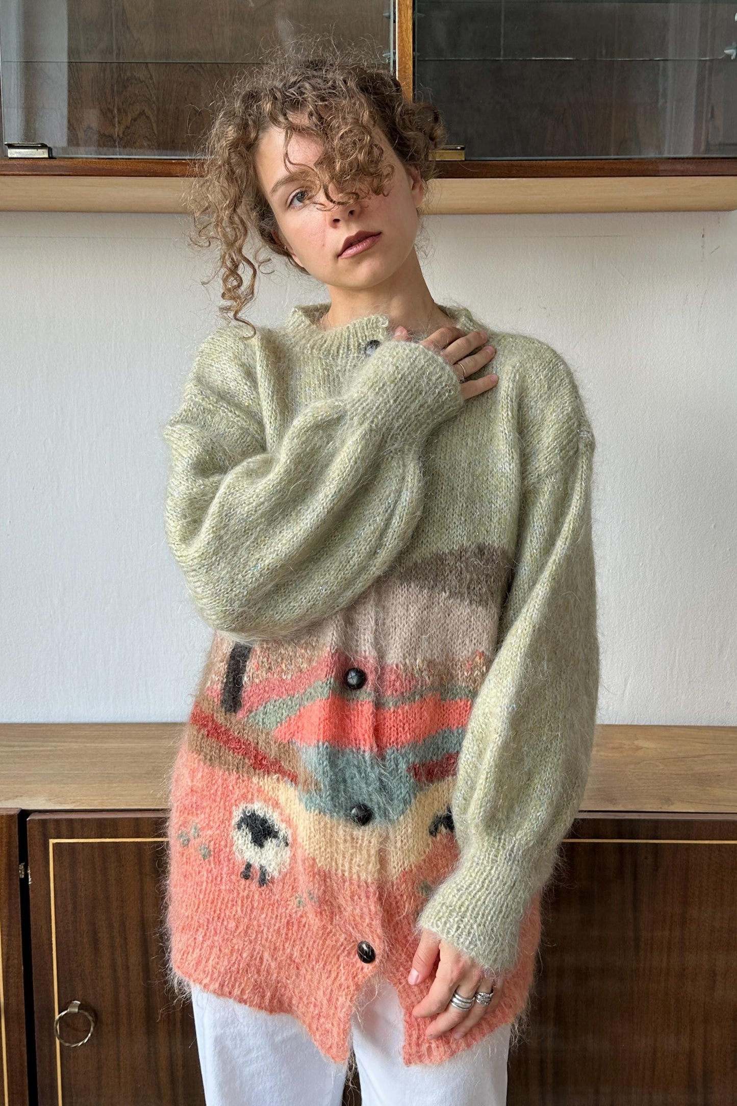 A young woman with curly hair wearing a colorful, patterned sweater stands in front of a wooden cabinet.