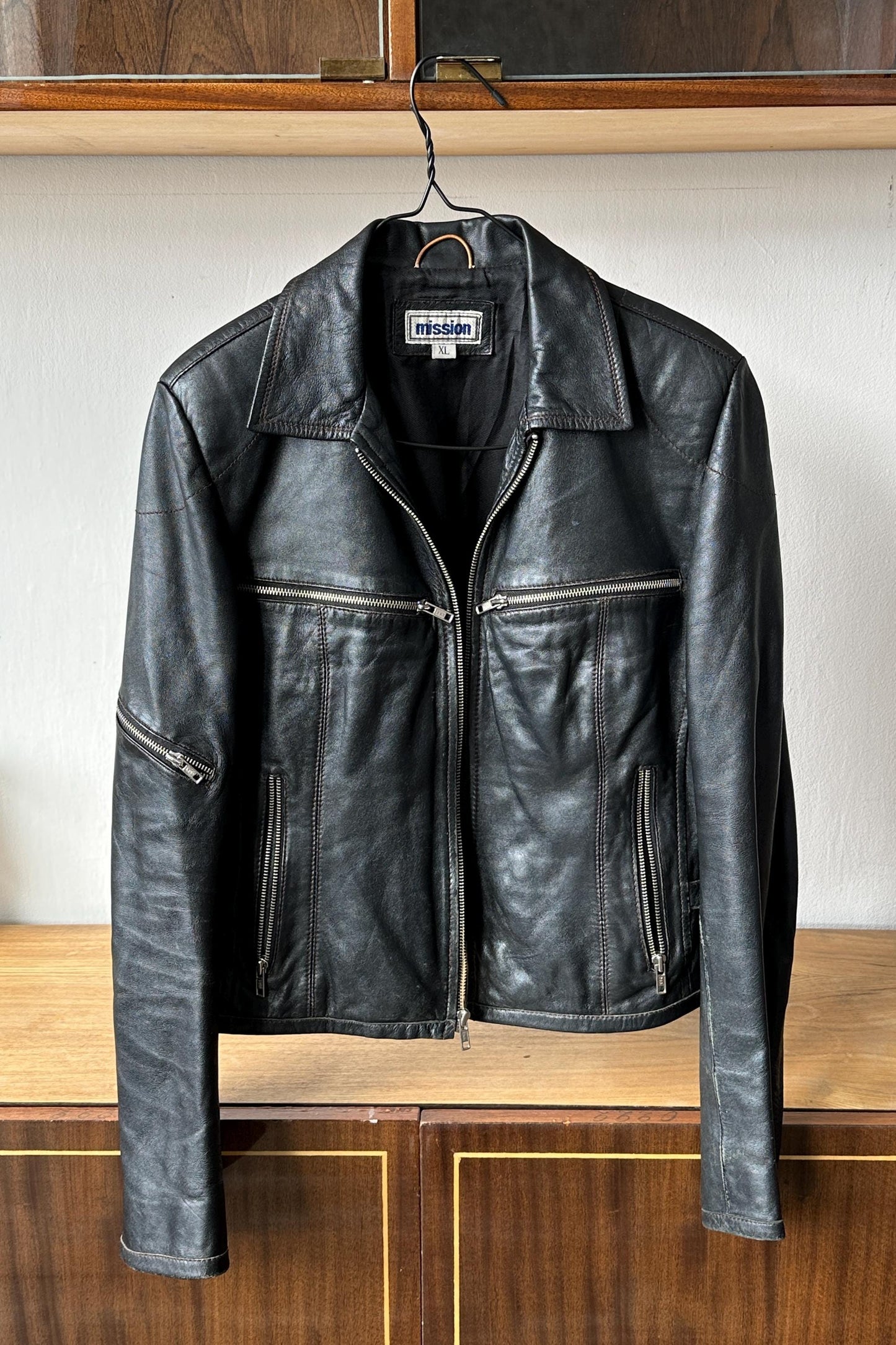 A black leather jacket with a collar and zipper is hanging on a metal hanger against a white wall.