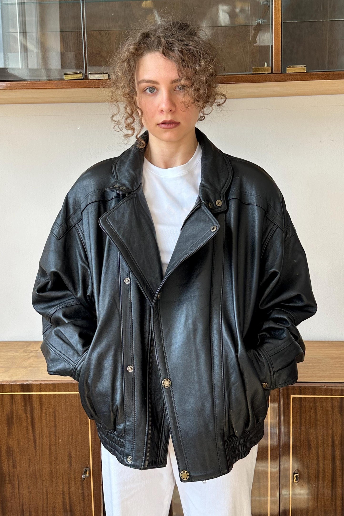A woman wearing a black leather jacket stands in front of a wooden cabinet.