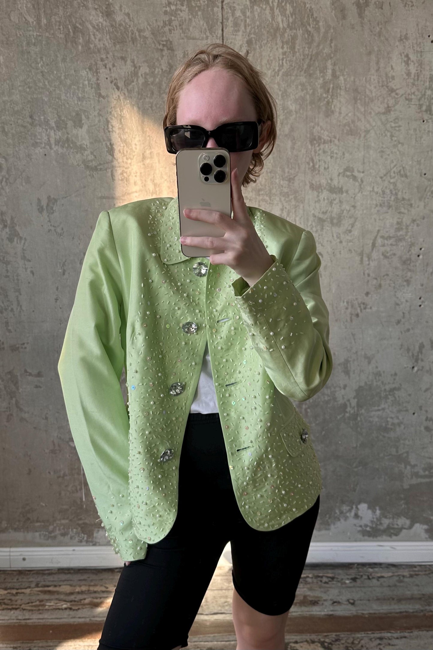 A person wearing a green jacket with sparkly details and sunglasses is taking a selfie with a smartphone.