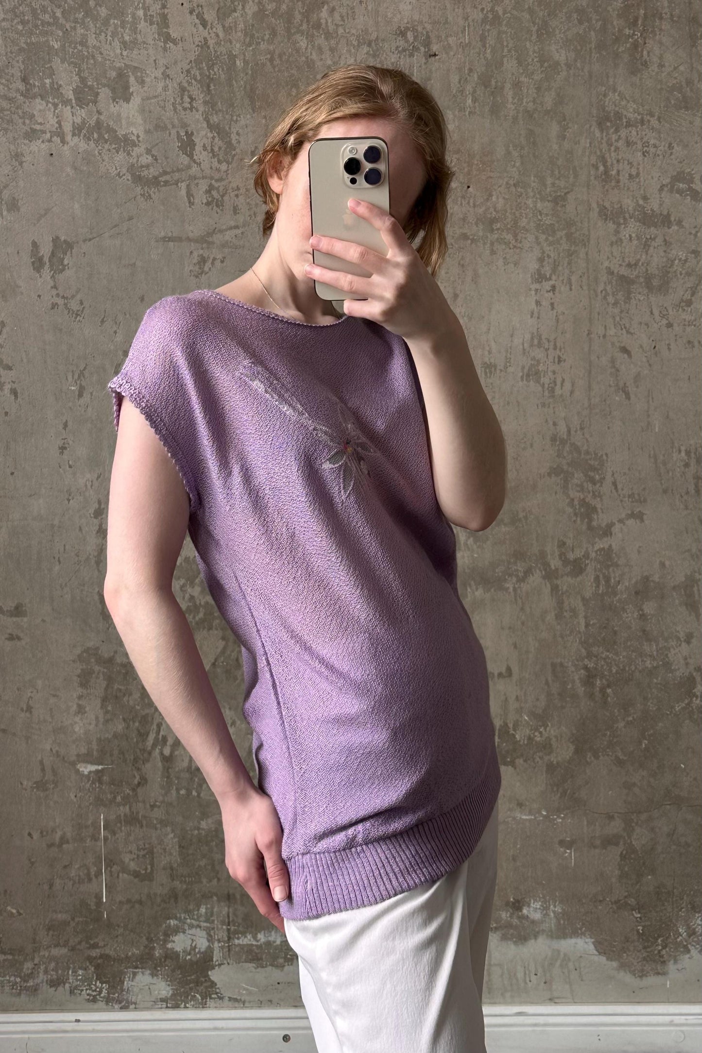A person is taking a selfie with a smartphone, wearing a purple top and white pants, against a textured concrete wall.