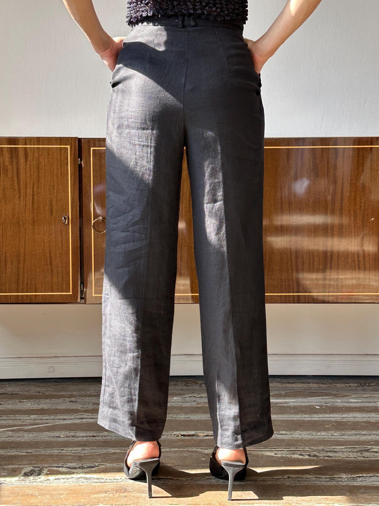 A person wearing gray pants and high heels stands with their hands in their pockets, facing away from the camera.