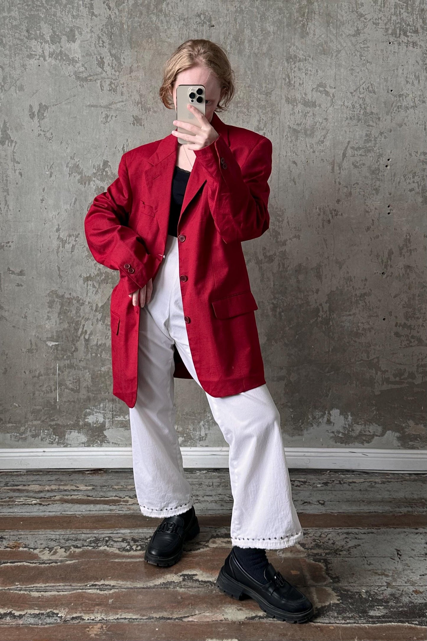 A person wearing a red jacket, white pants, and black shoes is taking a selfie with a smartphone against a concrete wall.