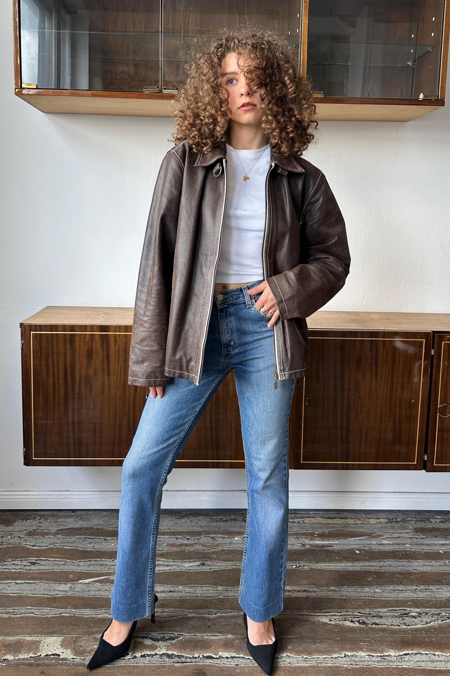 A woman with curly hair wearing a brown leather jacket, white shirt, and blue jeans stands in a room with wooden cabinets.