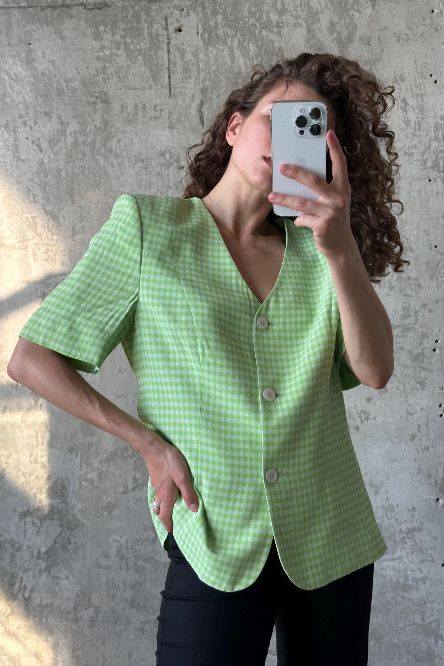 A woman with curly hair is taking a selfie with her phone, wearing a green and white checkered blazer.