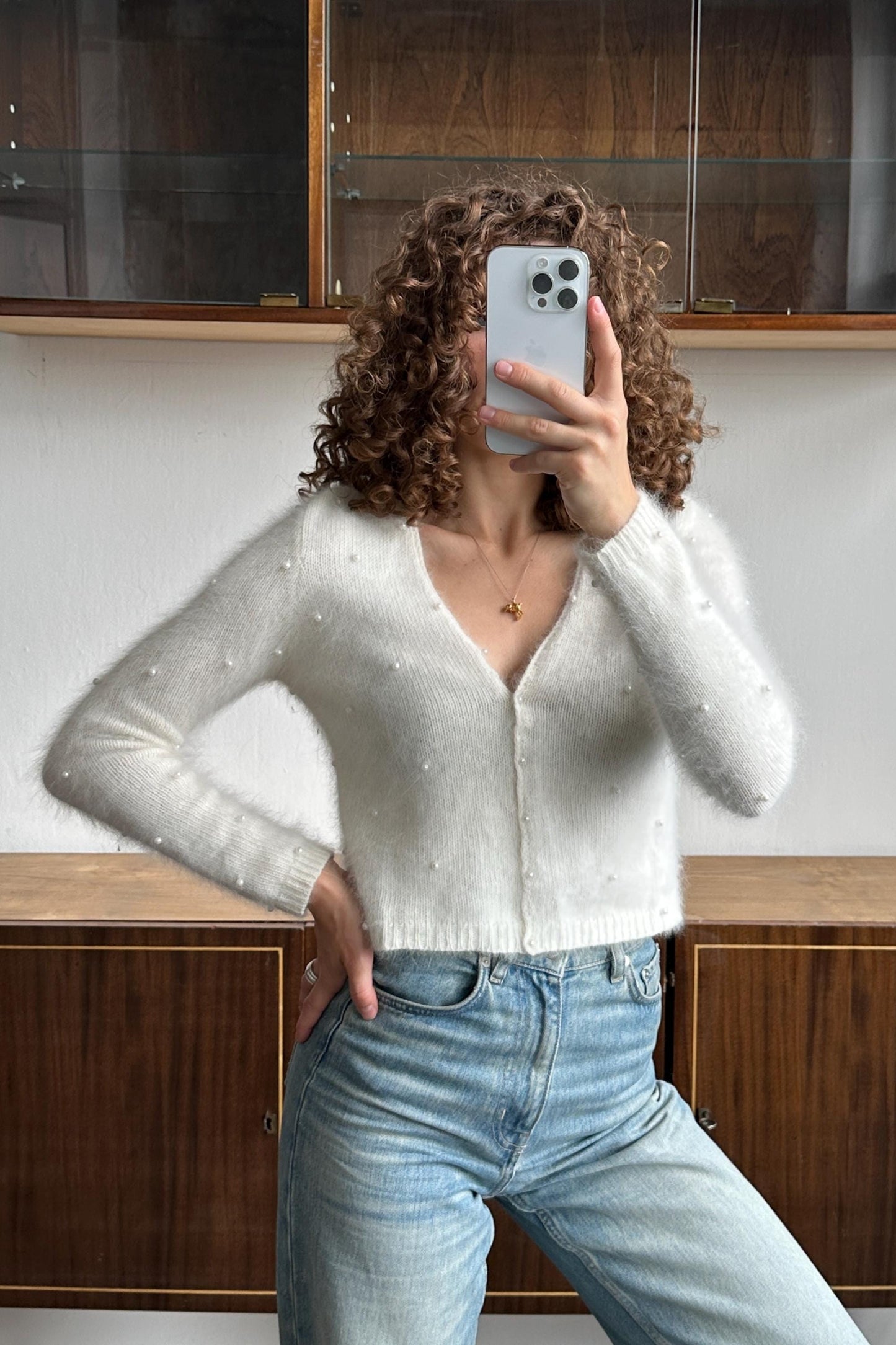 A woman with curly hair is taking a selfie with her iPhone, wearing a white sweater with polka dots and blue jeans.