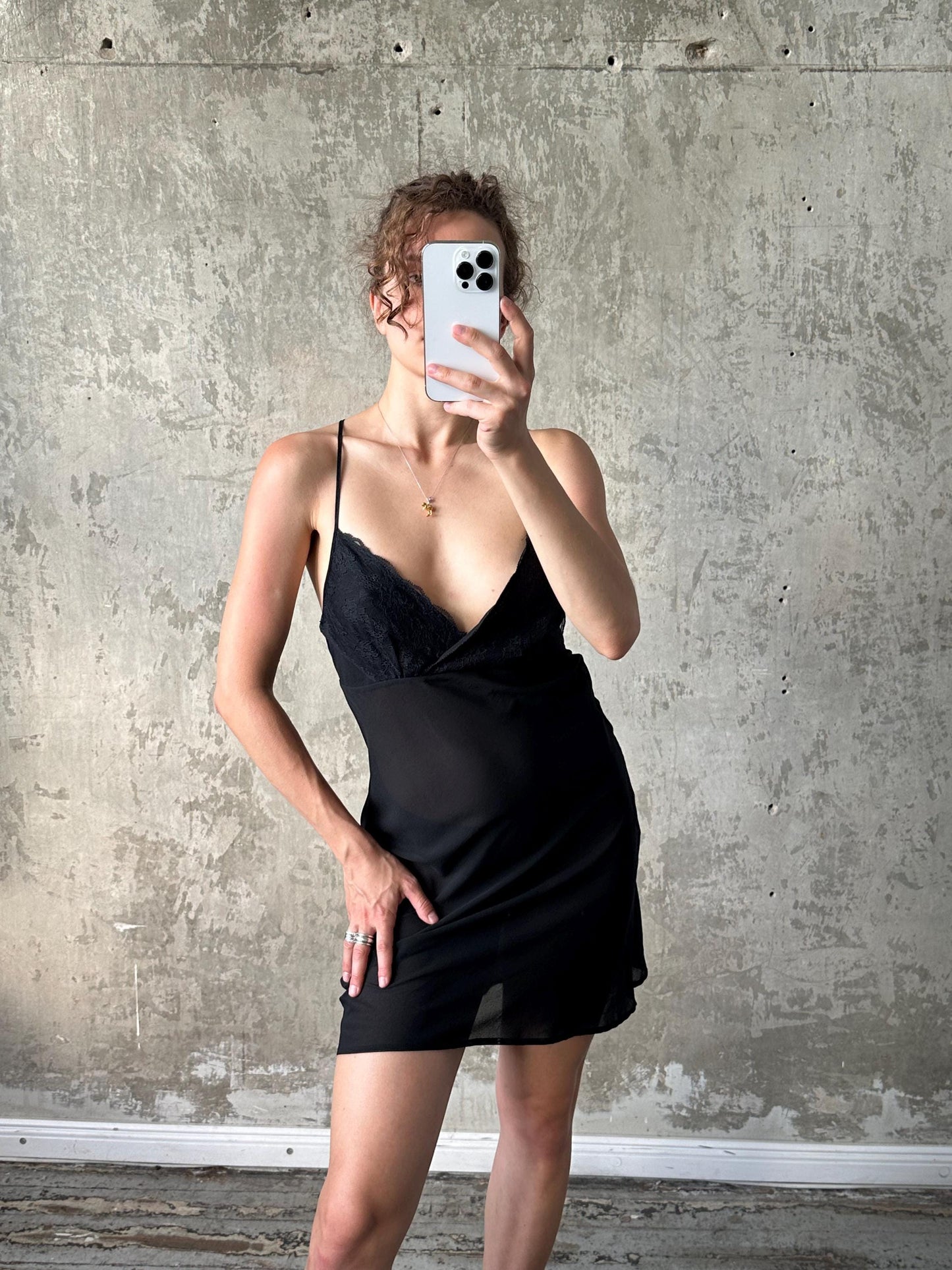 A woman in a black dress is taking a selfie with a white phone against a concrete wall.