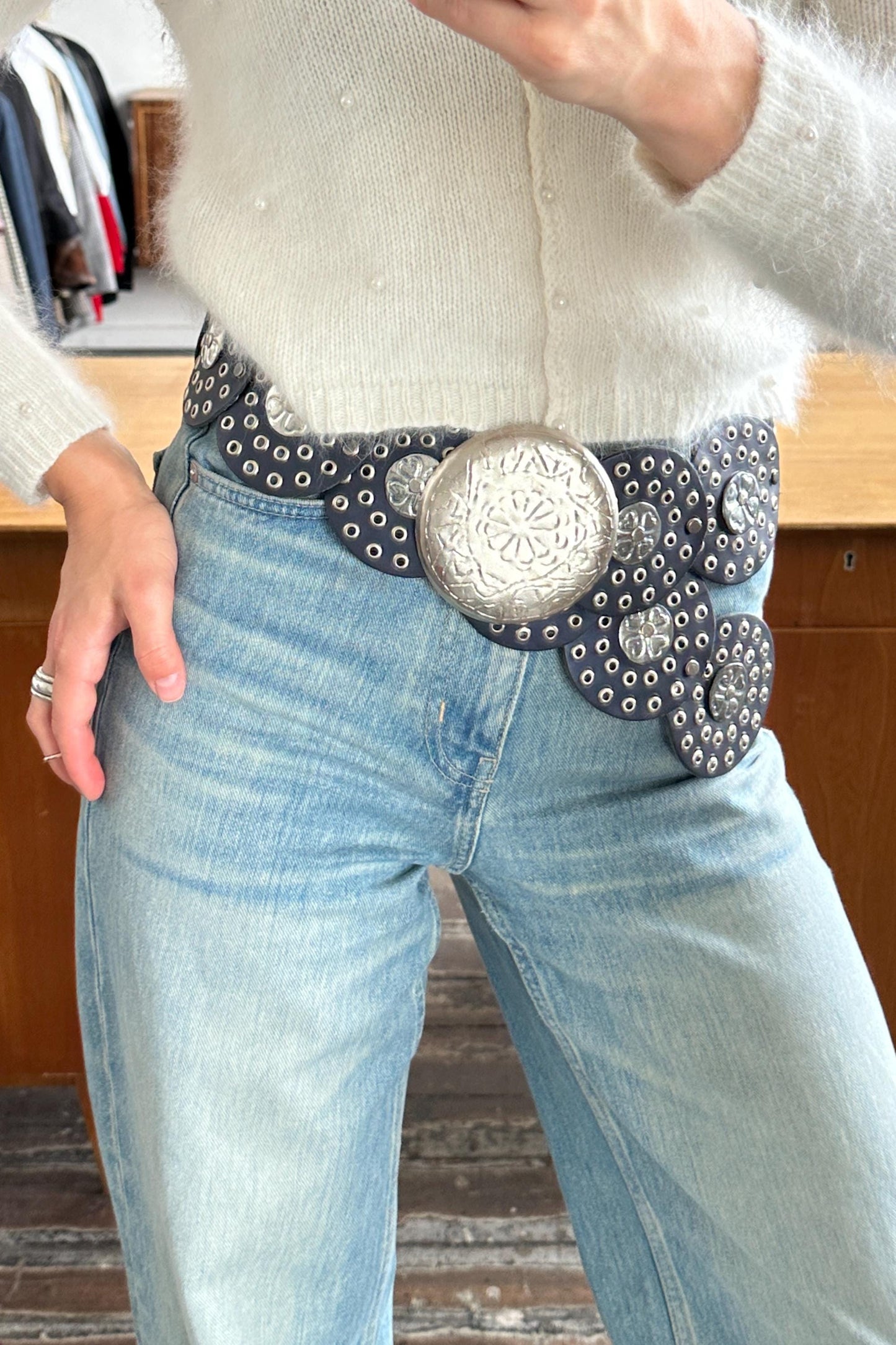 A person wearing a white sweater and blue jeans, with a large silver belt featuring a floral design.