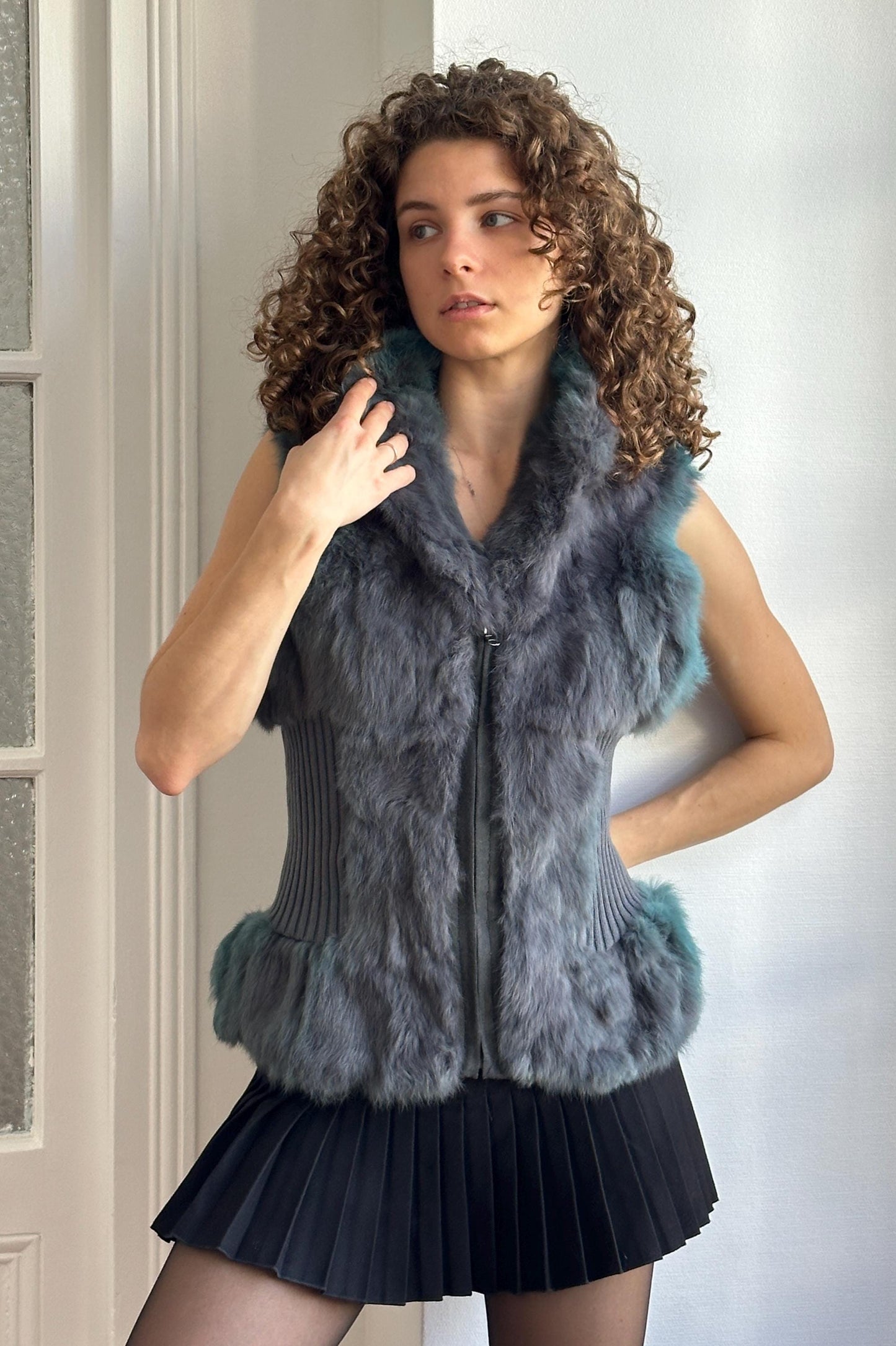 A woman with curly hair wearing a gray fur vest and a black skirt stands in front of a white wall.