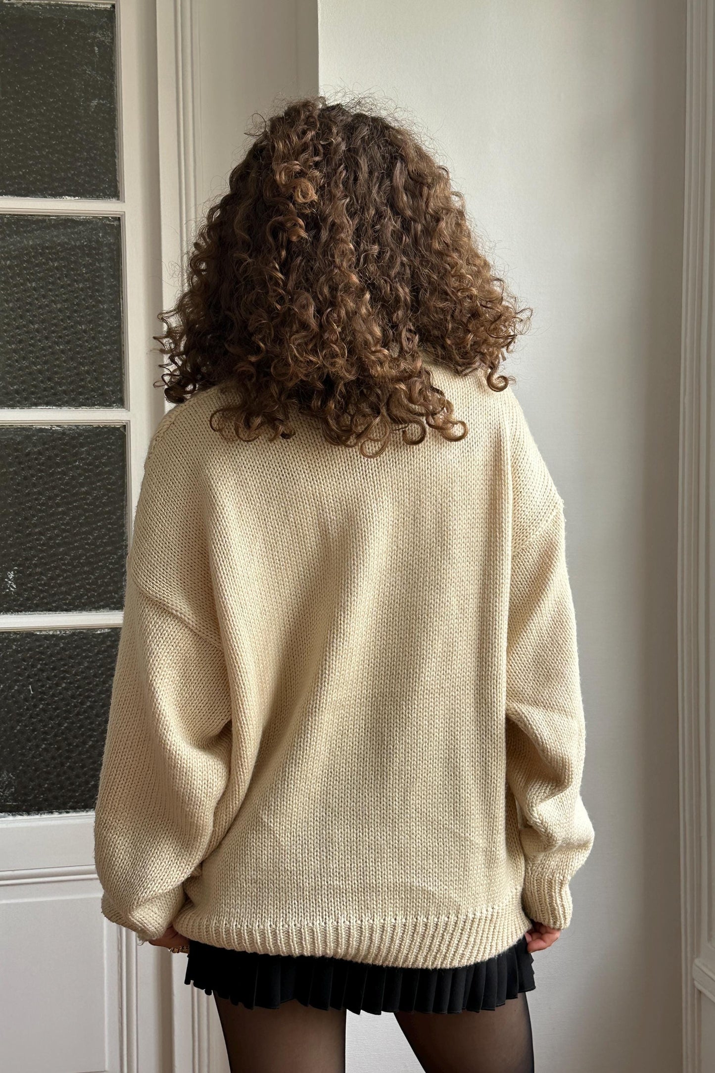 a person with curly hair wearing a cream-colored sweater and a black skirt, standing in front of a white door.