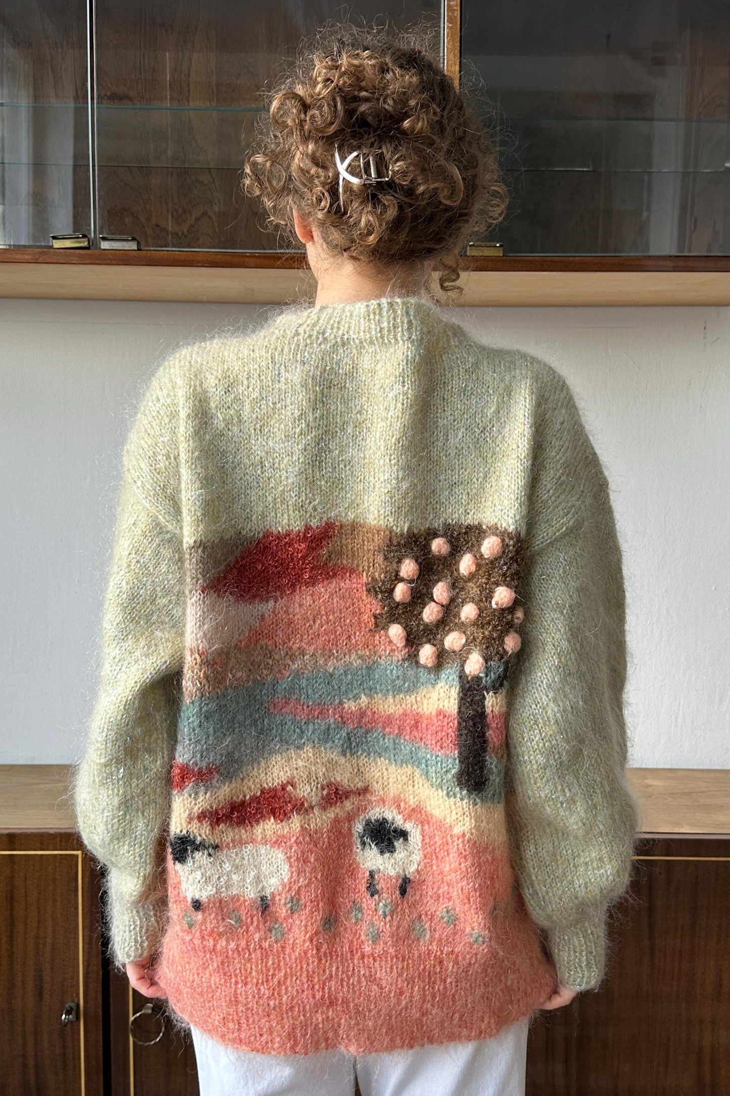 A person wearing a large, colorful knitted sweater with a pattern of sheep and a tree on the back.