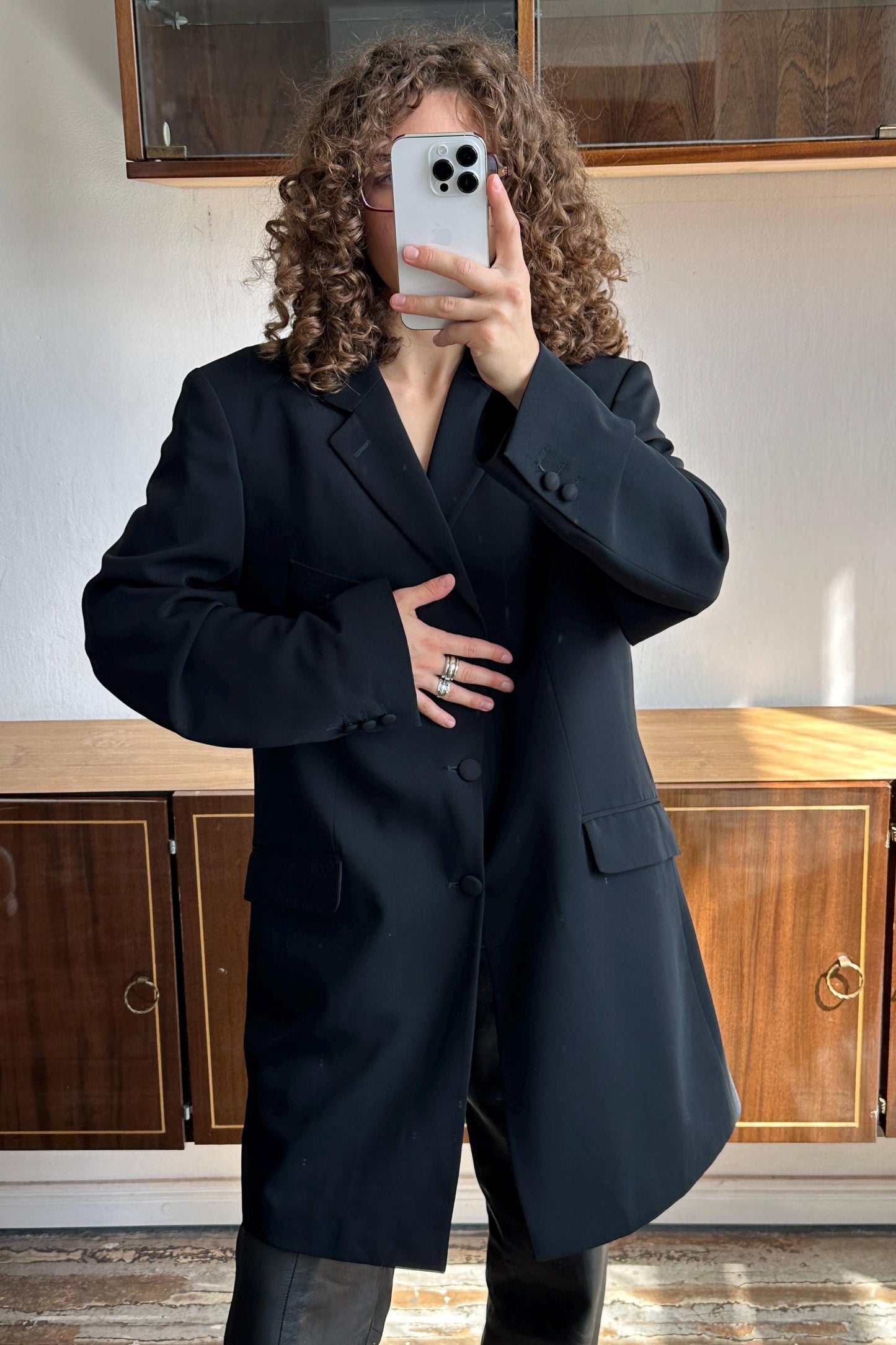 A person with curly hair is taking a selfie with a white phone, wearing a black blazer.