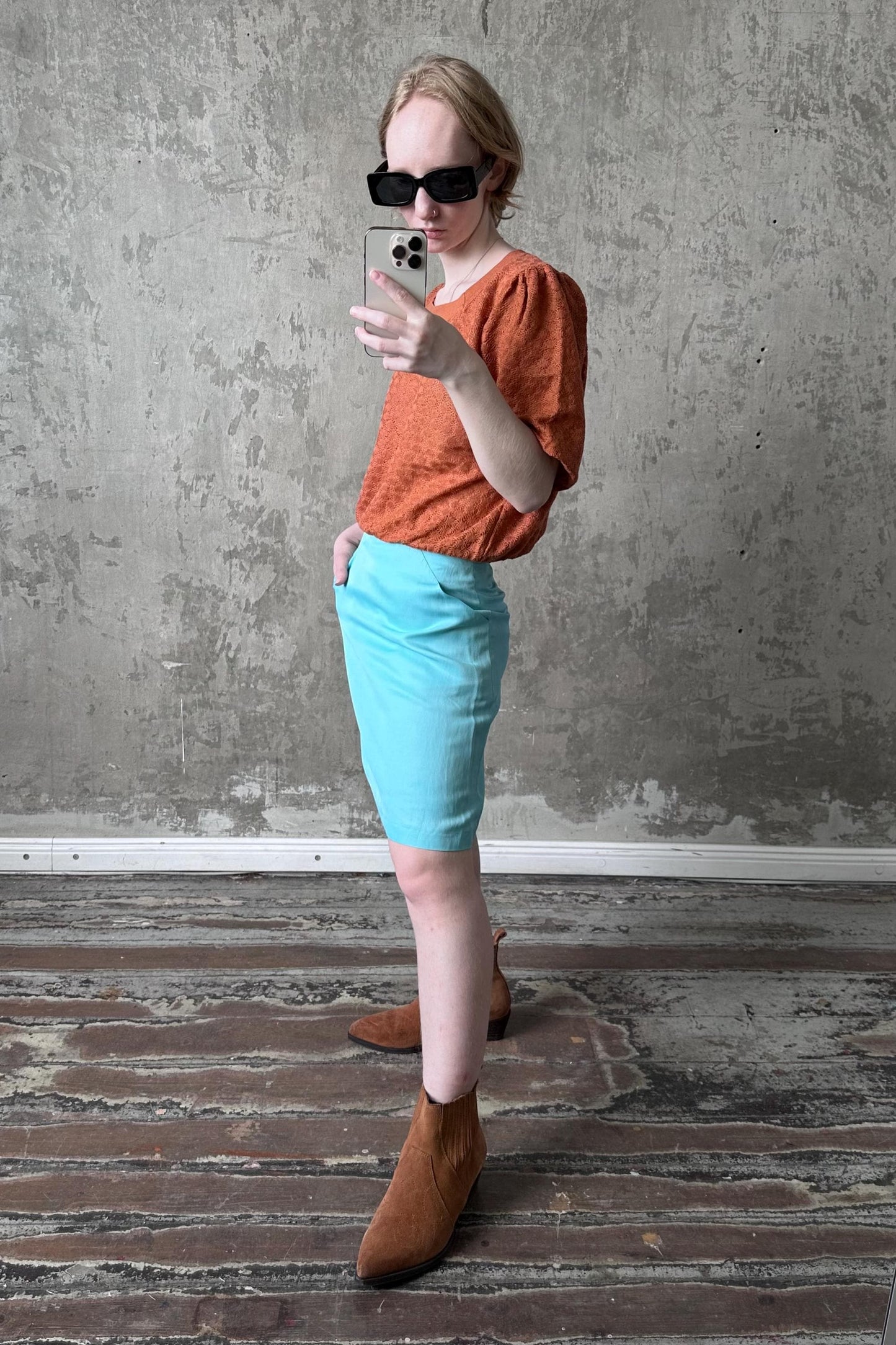 A person is taking a selfie with a smartphone, wearing a bright orange top, a light blue skirt, and brown boots, standing in front of a concrete wall.