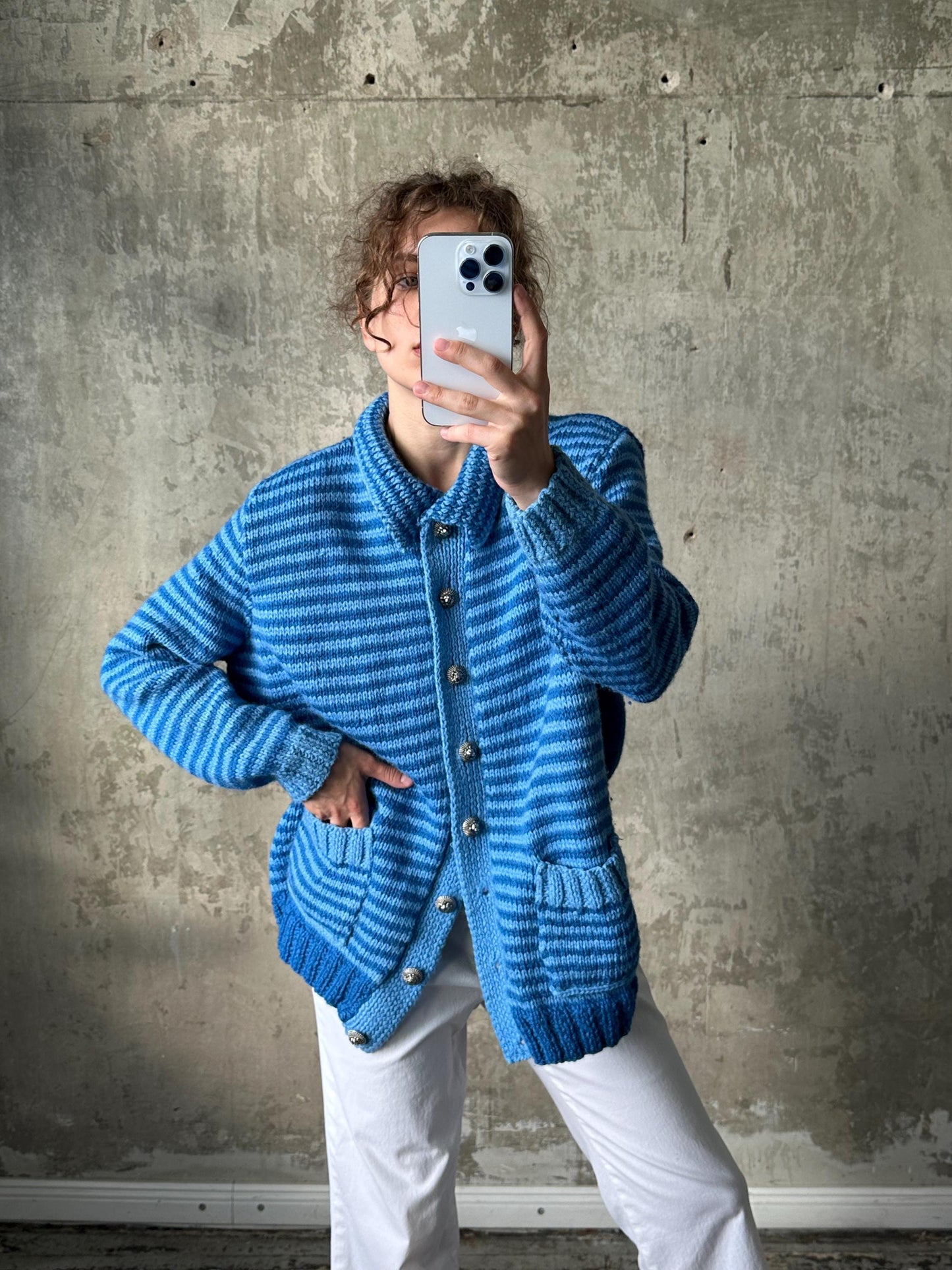 A person is taking a selfie with a smartphone, wearing a blue striped cardigan and white pants.