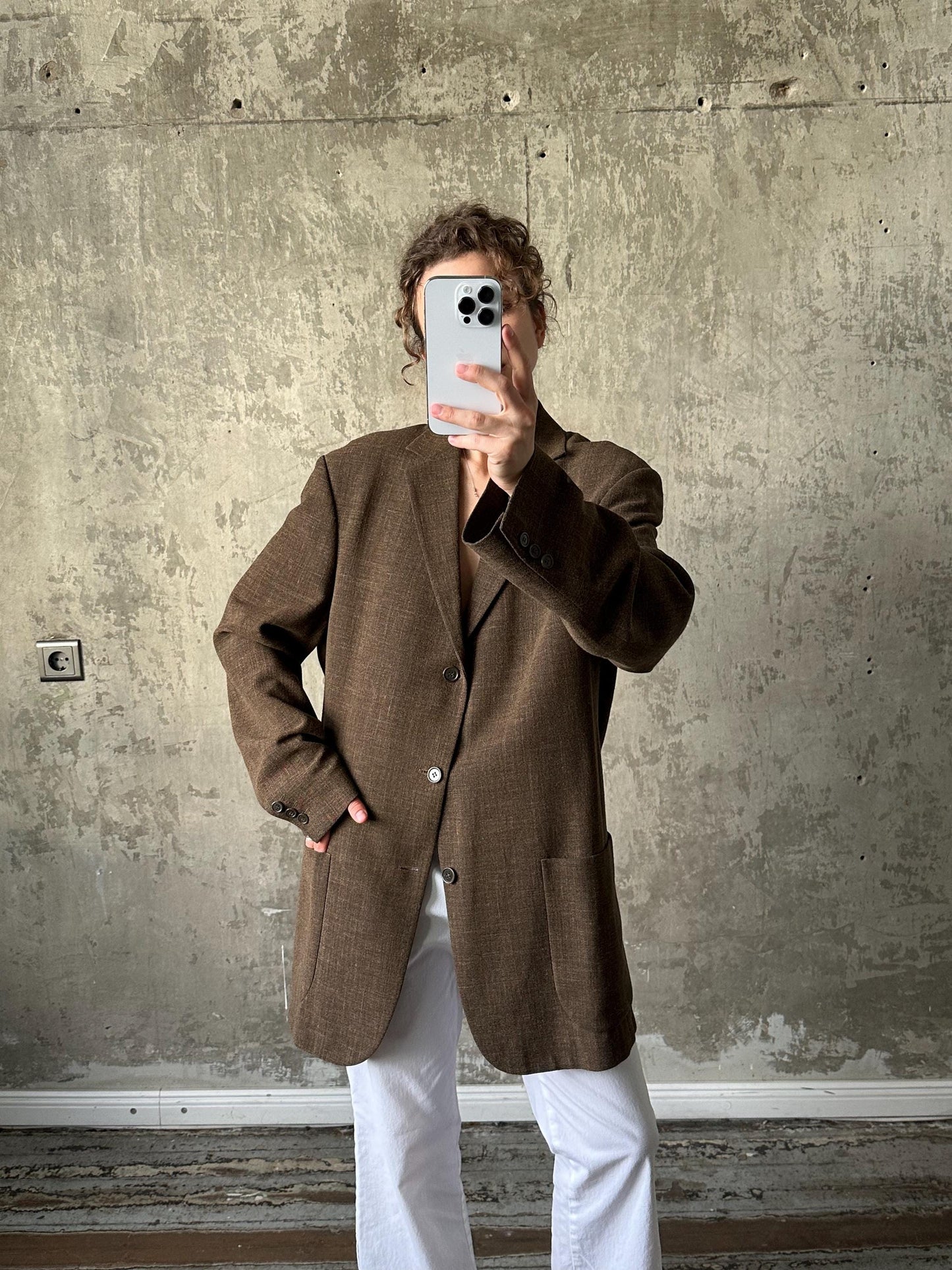A person is taking a selfie with a smartphone, wearing a brown blazer and white pants, against a concrete wall background.