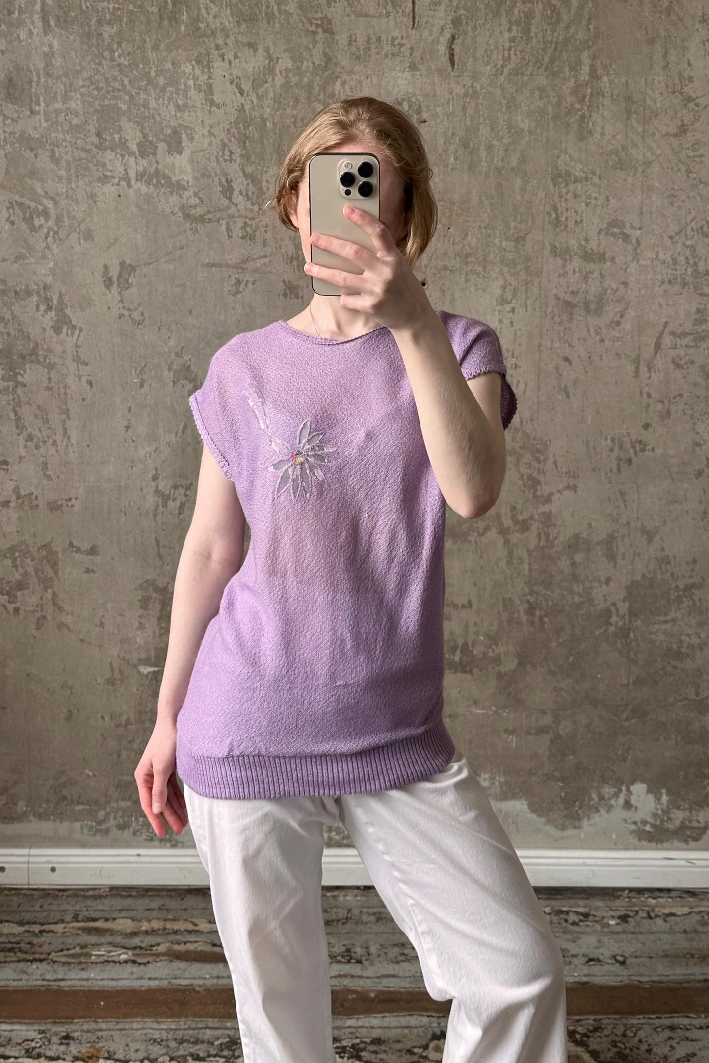 A person is taking a selfie with a smartphone, wearing a purple sweater with a flower design and white pants.