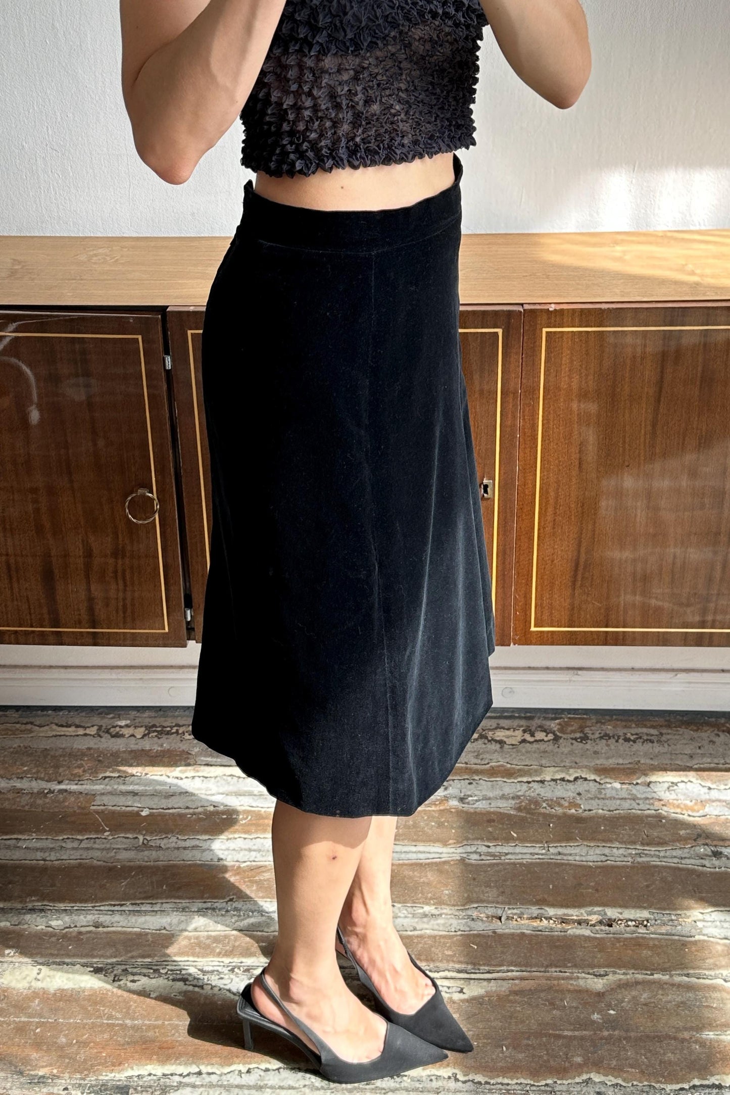 A woman wearing a black skirt and top stands in a room with wooden cabinets in the background.